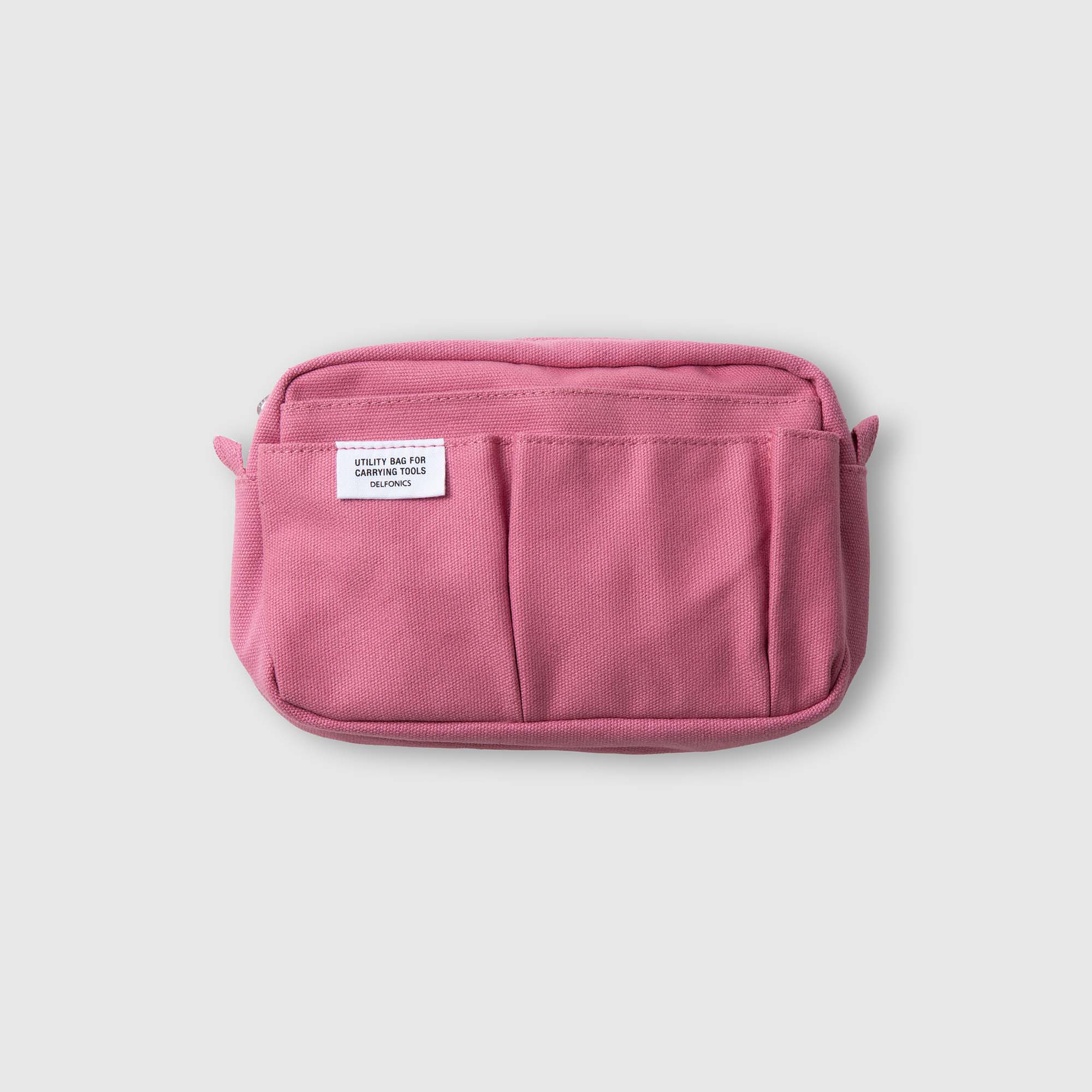 Eine kleine, rechteckige, rosa Delfonics-Innentasche „Inner Carrying Bag S“ aus Baumwolle mit zwei Vordertaschen und einem weißen Etikett oben links wird vor einem schlichten hellgrauen Hintergrund präsentiert – ideal als Organizer-Tasche oder Beutel für die Dinge des täglichen Bedarfs von Delfonics.