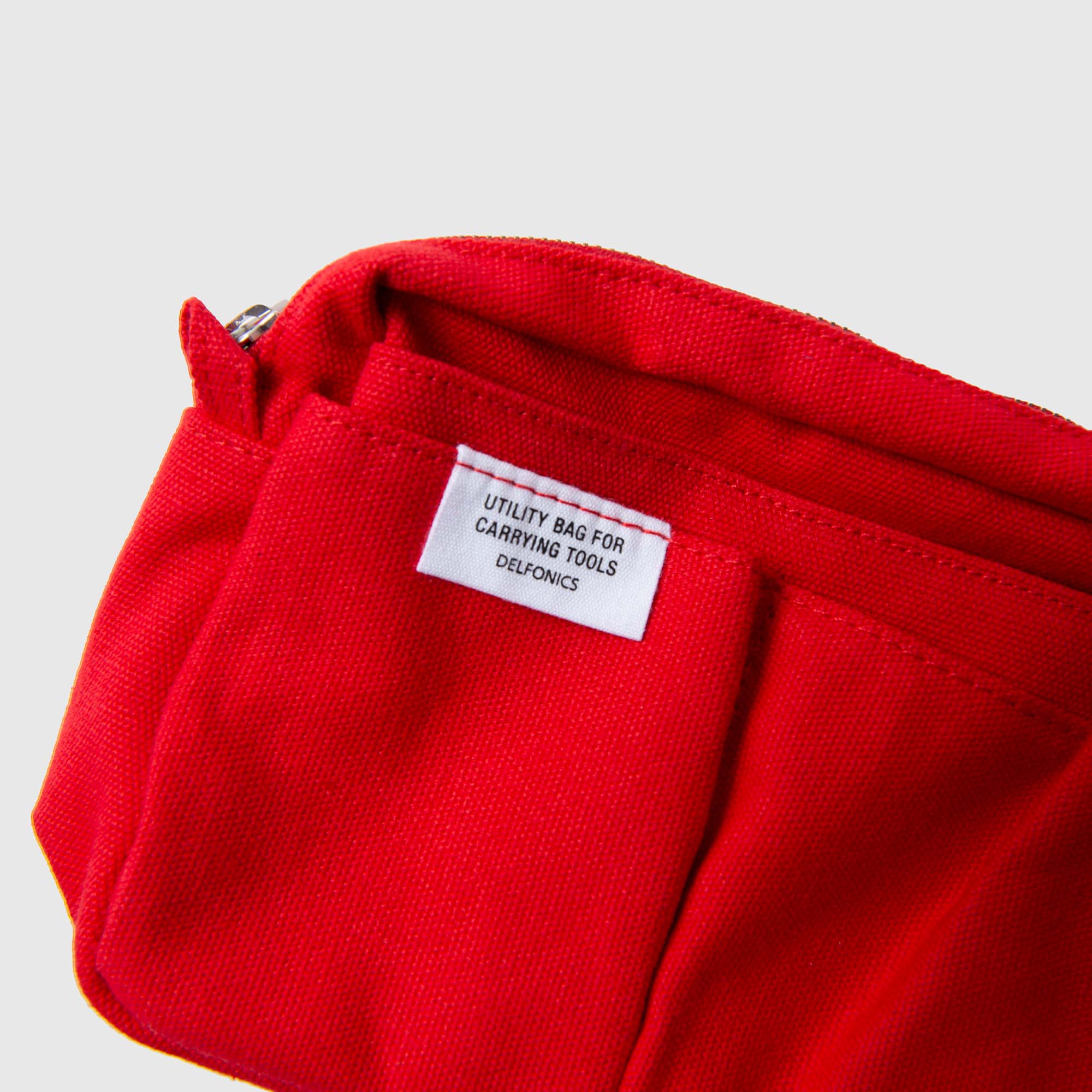 Eine Nahaufnahme der Delfonics Inner Carrying Bag S – einer roten Multifunktionstasche mit einem weißen Etikett, auf dem „Utility Bag for Carrying Tools DELFONICS“ steht – zeigt den Reißverschluss und die Details der Vordertasche. Diese vielseitige Tasche von Delfonics aus Baumwolle eignet sich perfekt, um Ihre wichtigsten Utensilien übersichtlich zu verstauen.