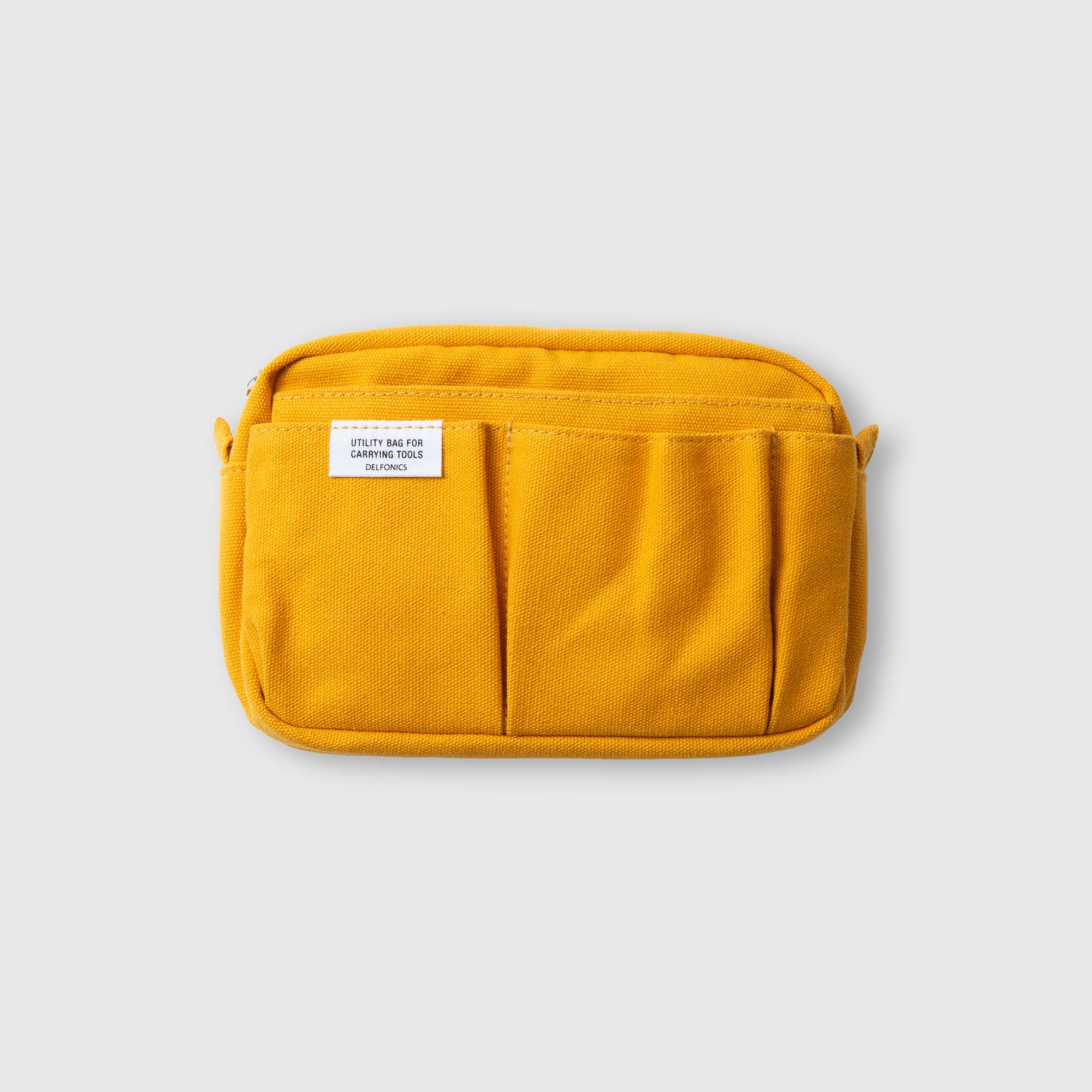 Eine kompakte, rechteckige, gelbe Stofftasche von Delfonics – die „Delfonics Inner Carrying Bag S“ – mit Fronttaschen und einem weißen Etikett oben links, dargestellt auf einem schlichten hellgrauen Hintergrund; ideal als Organizer-Tasche oder praktische Baumwolltasche für alltägliche Utensilien.
