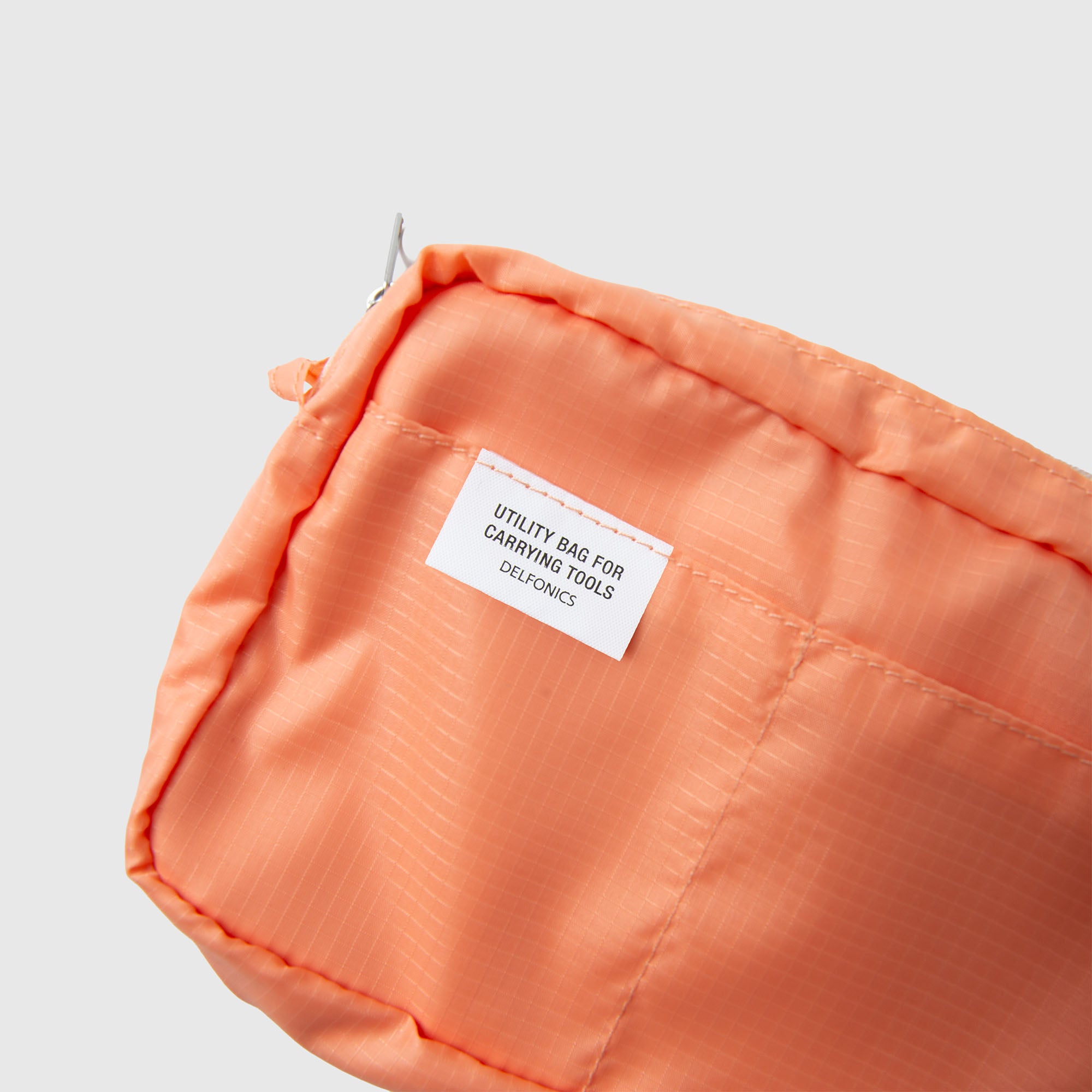 Nahaufnahme einer orangefarbenen Delfonics-Innentasche „AIR S“ aus strapazierfähigem Ripstop-Gewebe mit einem weißen Etikett, auf dem „Utility bag for carrying tools“ steht. DFLYGWQGLS vor einem schlichten hellgrauen Hintergrund. Diese leichte Tasche von Delfonics eignet sich ideal zum Ordnen kleiner Gegenstände oder zum Transportieren von wichtigen Utensilien wie einem Notizbuch im A6-Format.
