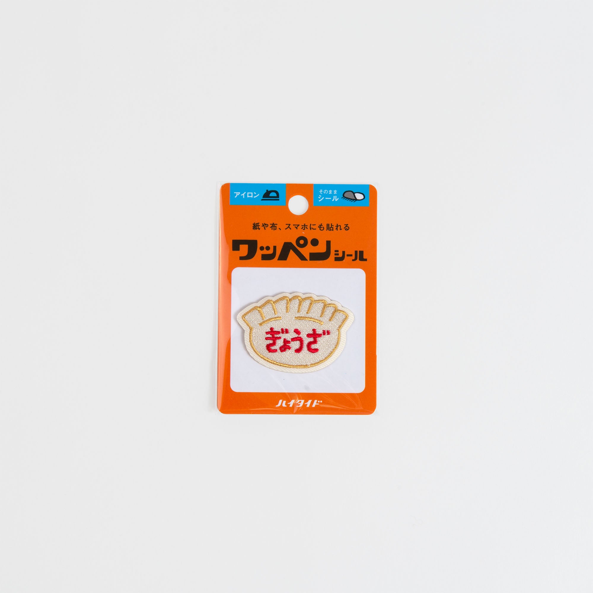 Der Hightide New Retro Patch Sticker von Hightide ist ein knödelförmiger, gestickter Aufnäher zum Aufbügeln mit japanischem Text auf dem Aufnäher und seiner orangefarbenen Verpackung, der den Charme der Showa-Ära einfängt - perfekt als Retro-Accessoire, dargestellt auf weißem Hintergrund.