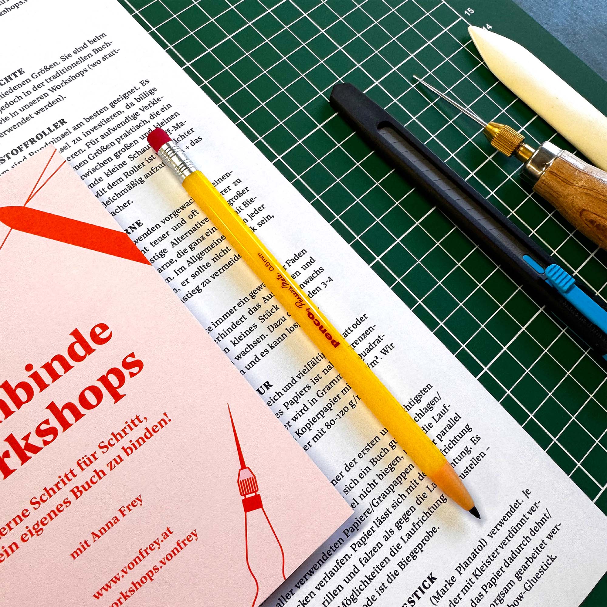 Ein gelber Bleistift, Buchbindewerkzeuge und Drucksachen liegen auf einer grünen Schneidematte. Ein rotes Heftchen weist auf den Workshop Crémant und Notizbuch von paperlux.store hin - ideal zum Erlernen von Notizbuchbindetechniken.