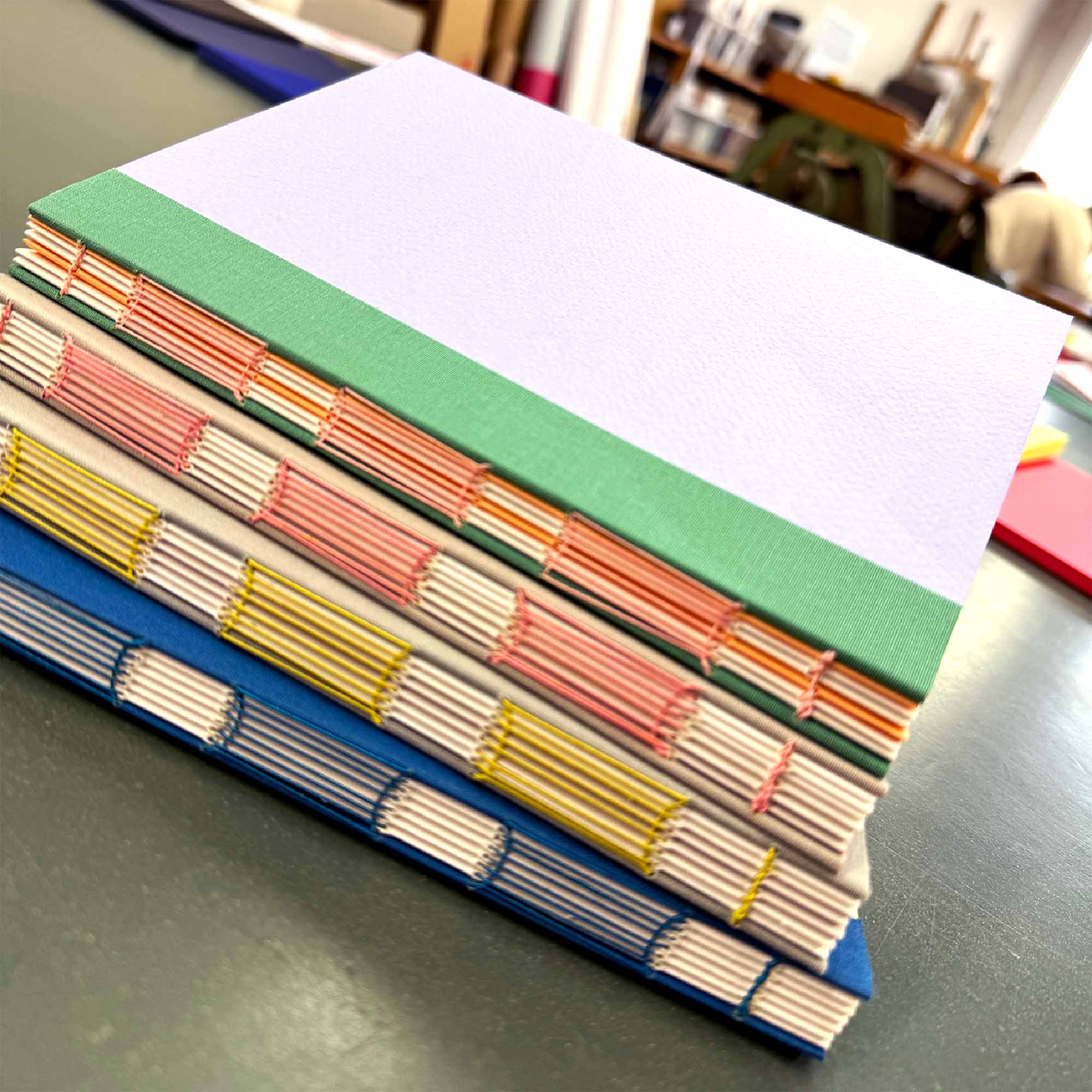 Eine Nahaufnahme von handgefertigten Büchern mit bunten Fadeneinbänden in Grün, Orange, Gelb und Blau aus dem Workshop Crémant und Notizbuch von paperlux.store, gestapelt auf einem Tisch in einem hellen, organisierten Raum.