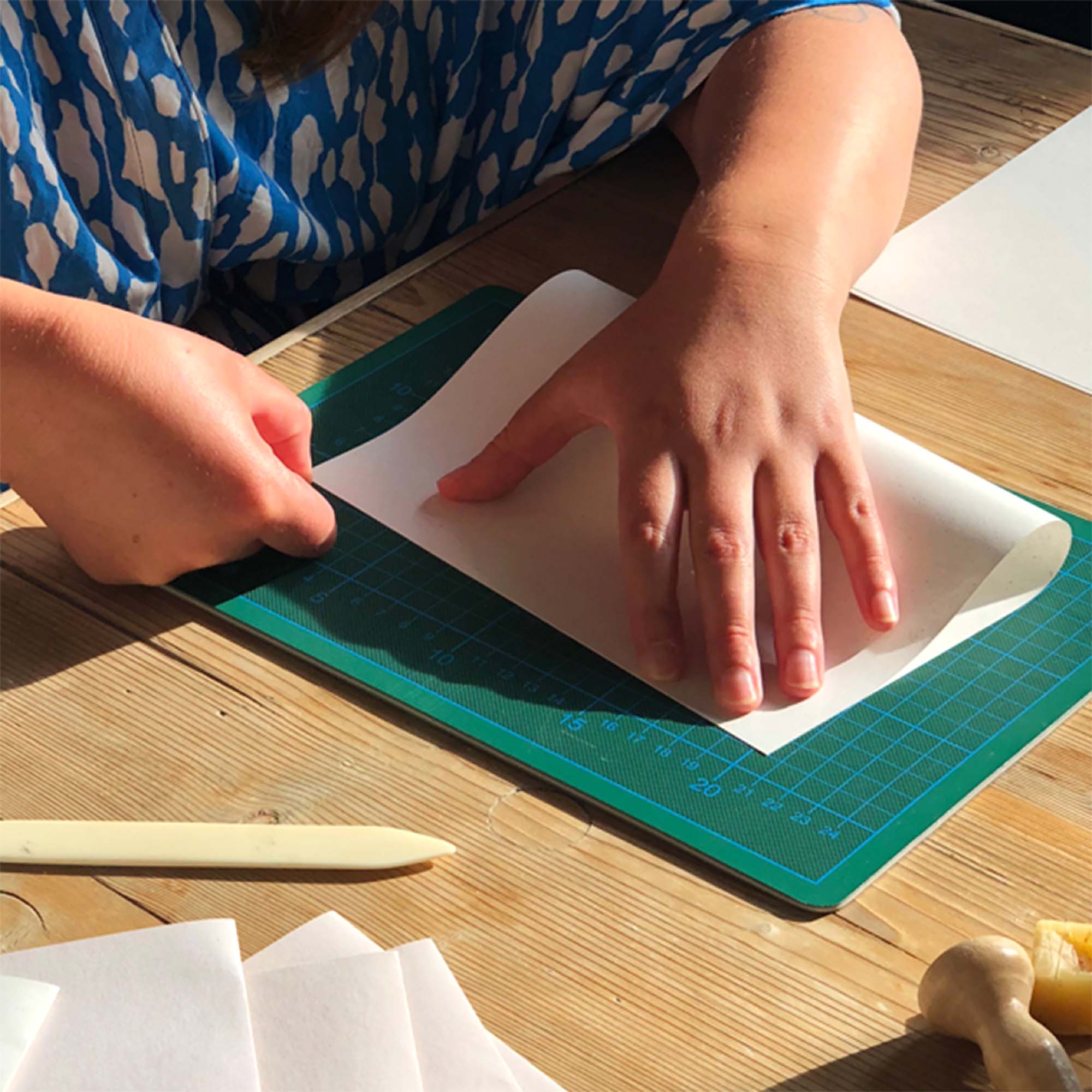 Im Workshop Crémant und Notizbuch von paperlux.store wird ein weißes Blatt Papier mit einem Knochenfalter auf einer grünen Schneidematte gefaltet; weitere Bastelwerkzeuge und Papiere liegen auf dem sonnenbeschienenen Holztisch.