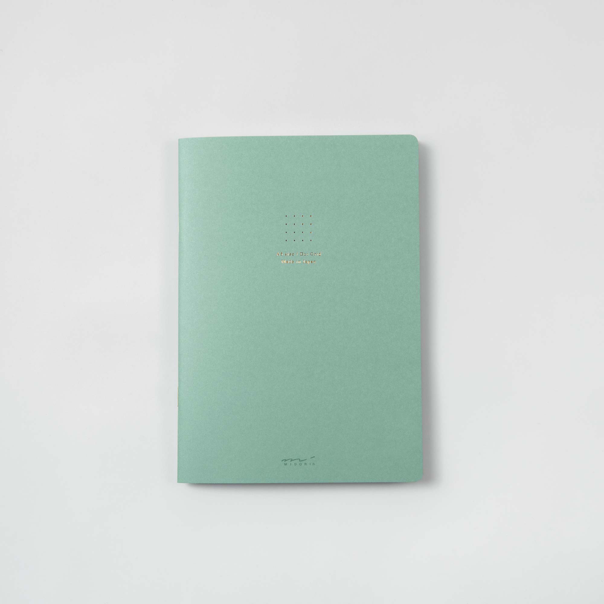 Auf einer weißen Oberfläche liegt ein geschlossenes Softcover-Notizbuch „Midori Notizhefte Color Dot Grid“ in pastellfarbenem Hellgrün und weist in der Mitte ein kleines, goldgeprägtes Design und Text auf.