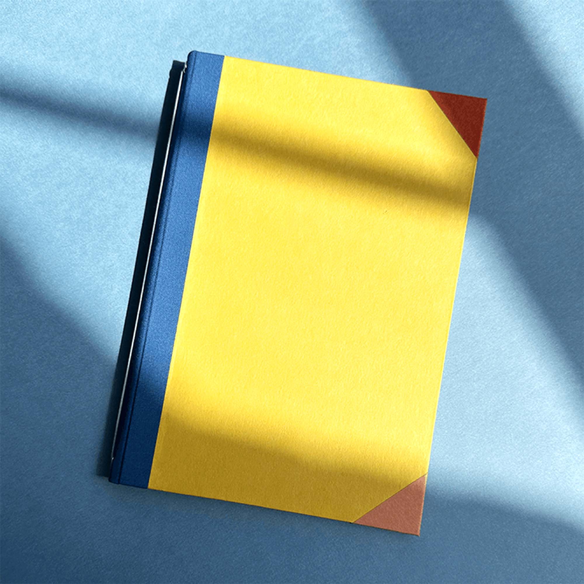 Ein geschlossenes gelbes Notizbuch mit blauem Rücken und braunen Ecken liegt auf einer hellblauen Fläche und fängt den kreativen Geist des Workshops Modulares Notizbuch von paperlux.store ein.