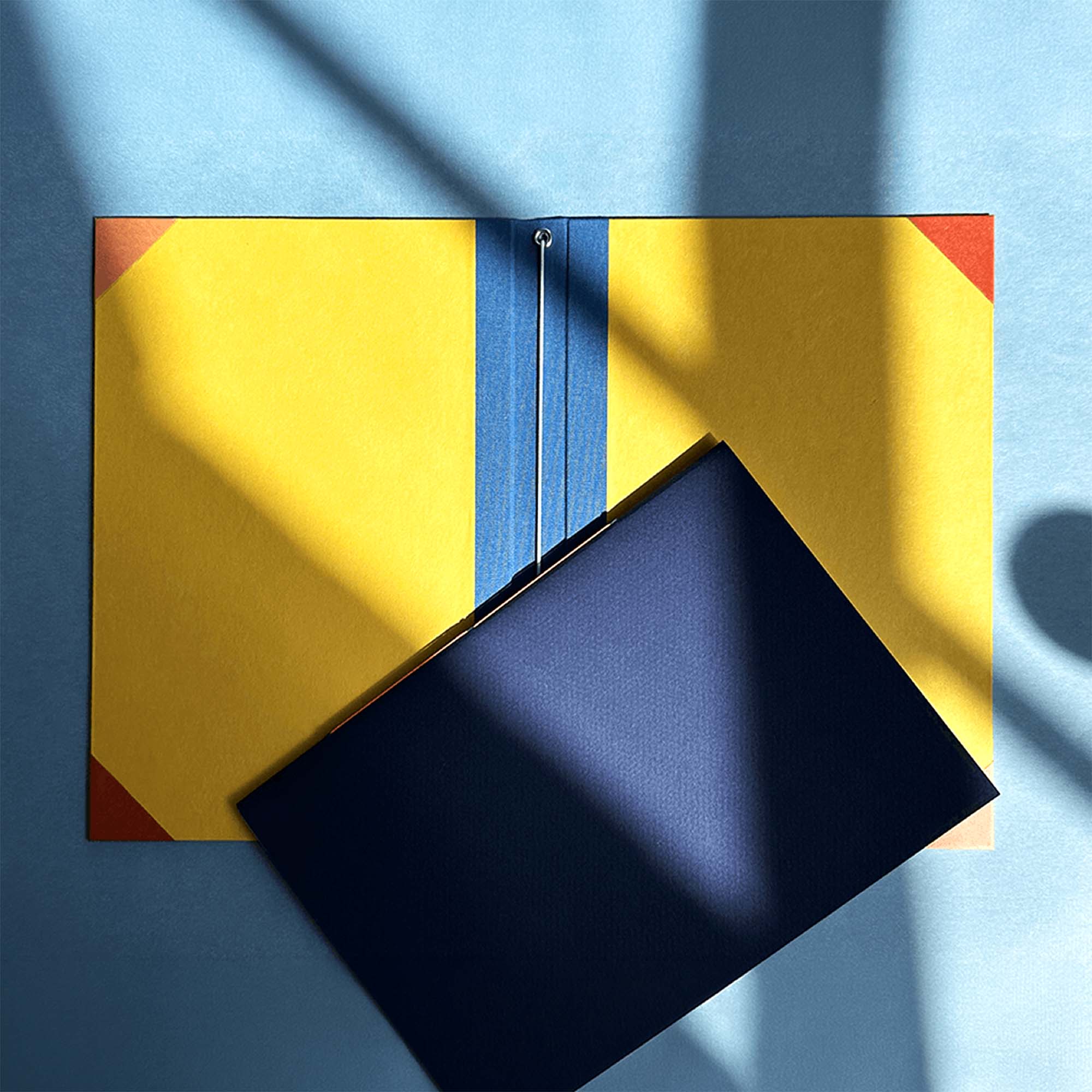 Der Workshop Modulares Notizbuch von paperlux.store zeigt einen gelben Einband mit roten Ecken und blauem Rücken, der aufgeschlagen auf einer blauen Fläche liegt, mit einem dunkelblauen Dokument im Inneren und Fensterschatten auf dem wiederverwendbaren Notizbuchdeckel.