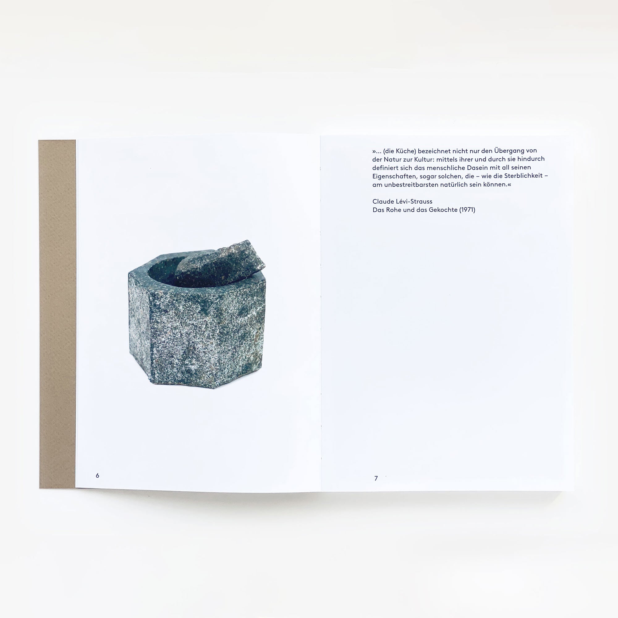 Ein aufgeschlagenes Buch mit dem Titel „Man kann keine Steine essen: Kochbuch eines japanischen Bildhauers“ von Shinroku Shimokawa zeigt die Seiten 6 und 7, mit einer grauen Steinschale aus regionalen Zutaten auf der linken Seite und einem deutschen Text, der mit „Kein König beeichtet schneller...“ beginnt, auf der rechten Seite. Der Hintergrund ist weiß. Marke: Prima Publikationen.