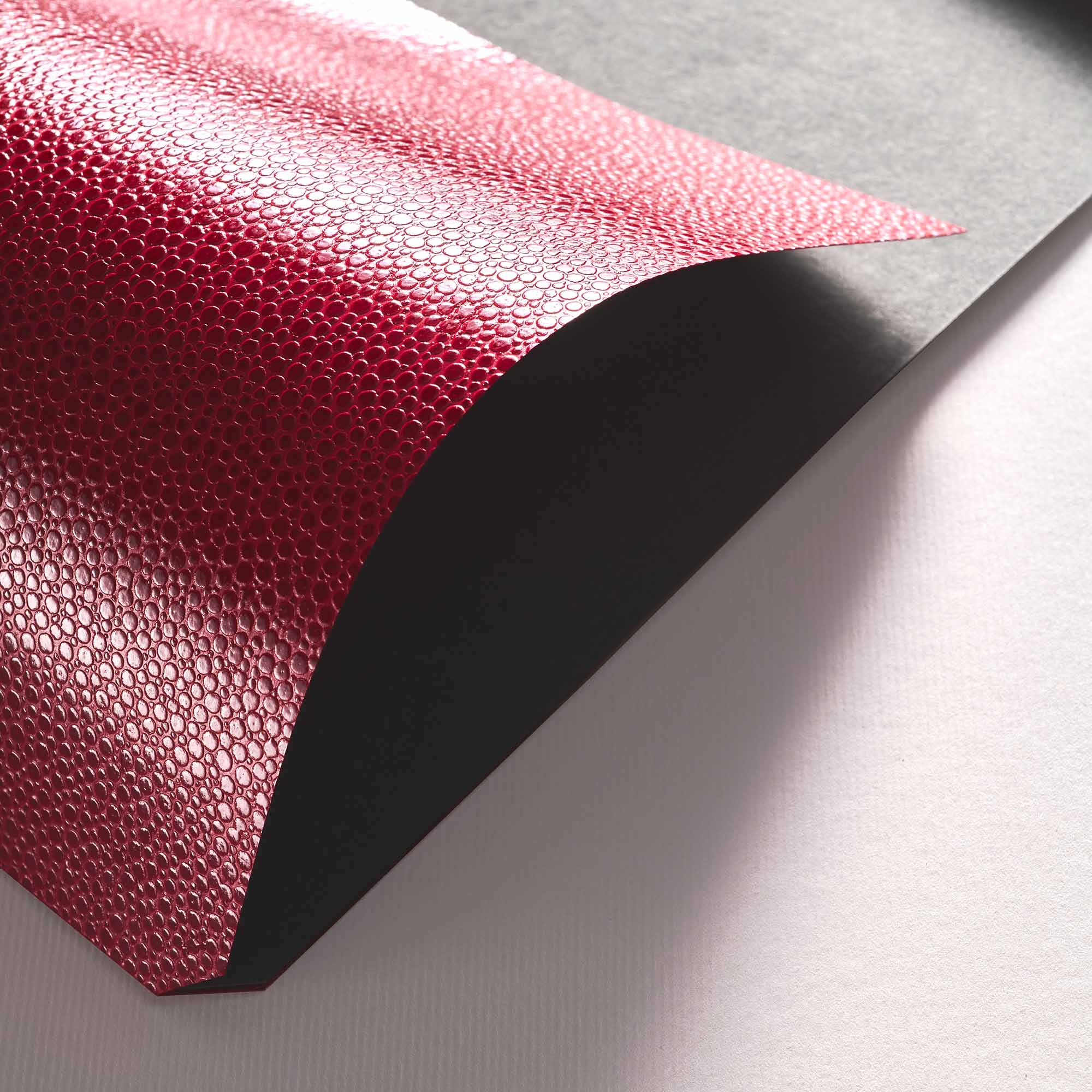 Eine Nahaufnahme eines schwarzen Blattes und eines glänzenden, strukturierten roten Blattes B96 Red Lipstick mit einem kleinen kreisförmigen Muster von paperlux.store, die beide auf einer weißen Fläche liegen.