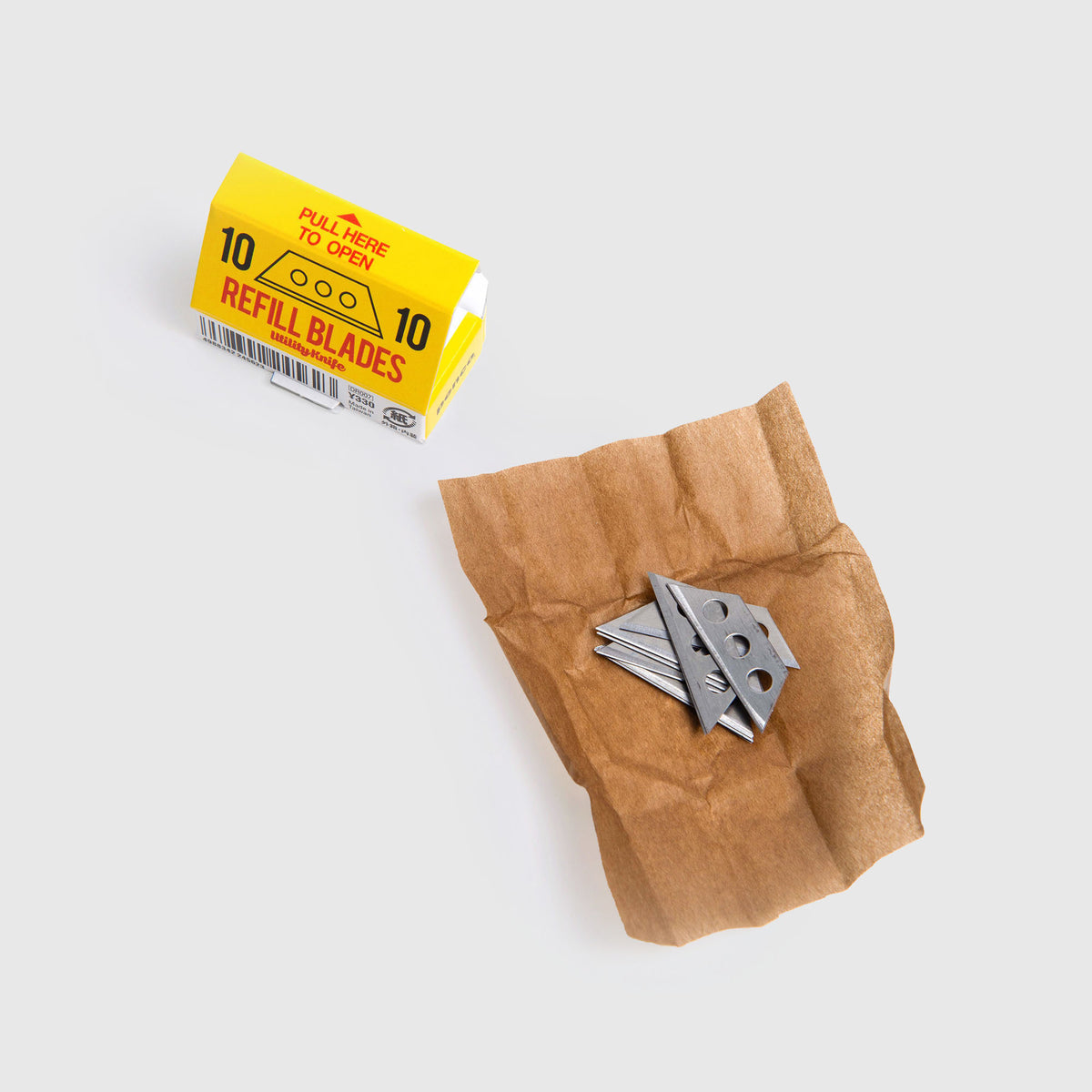 Eine kleine gelbe Schachtel mit der Aufschrift „REFILL BLADES“ von Penco’s Ersatzklingen: Cutter an der Kette steht offen neben mehreren losen Metallklingen, die auf einem Stück braunem Papier liegen, alles vor einem schlichten weißen Hintergrund.