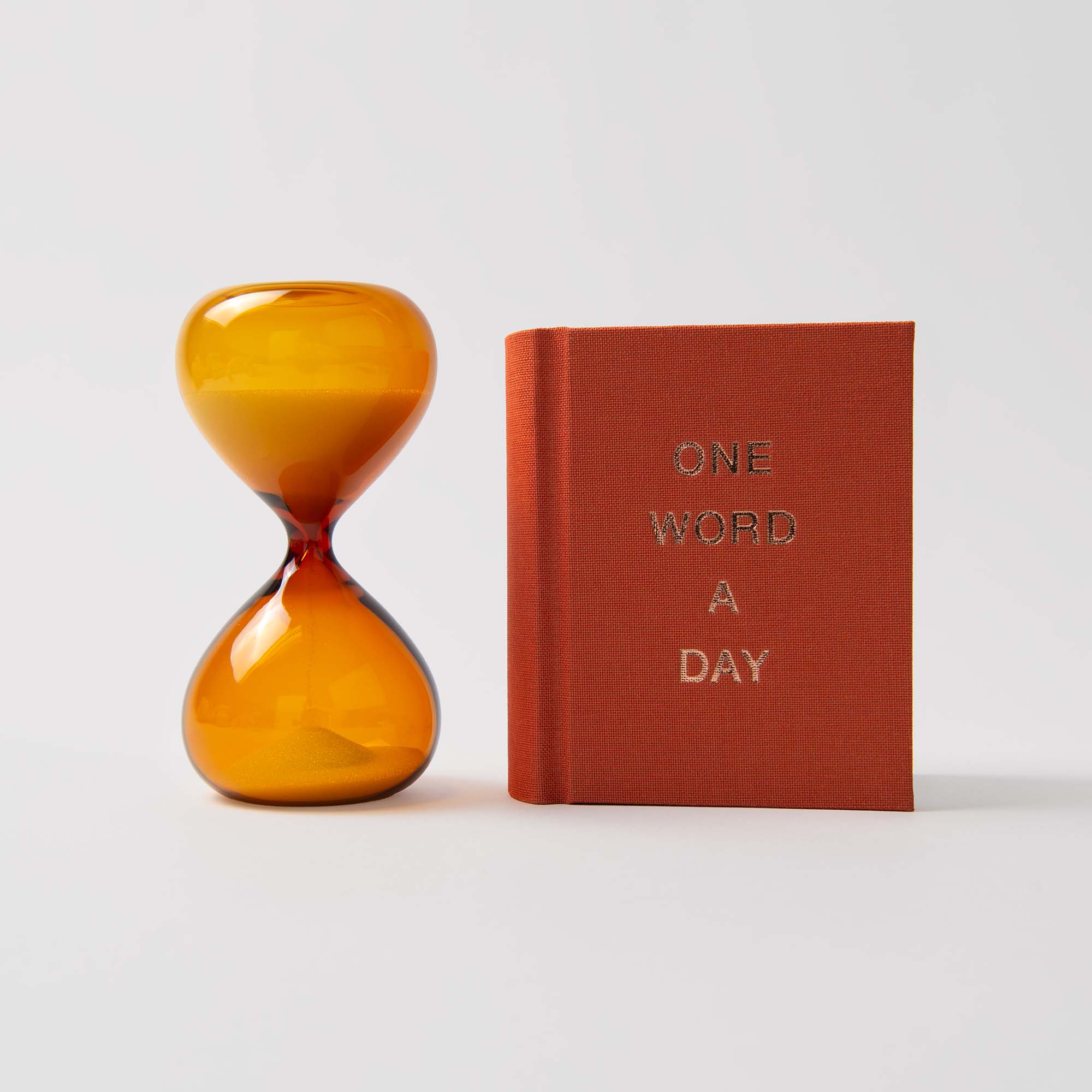 „One Word a Day“ mit Sanduhr von paperlux.store mit einem Timer aus Bernsteinglas und einem orangefarbenen Buch mit goldener Schrift ist ein nettes Geschenk vor einem schlichten weißen Hintergrund.