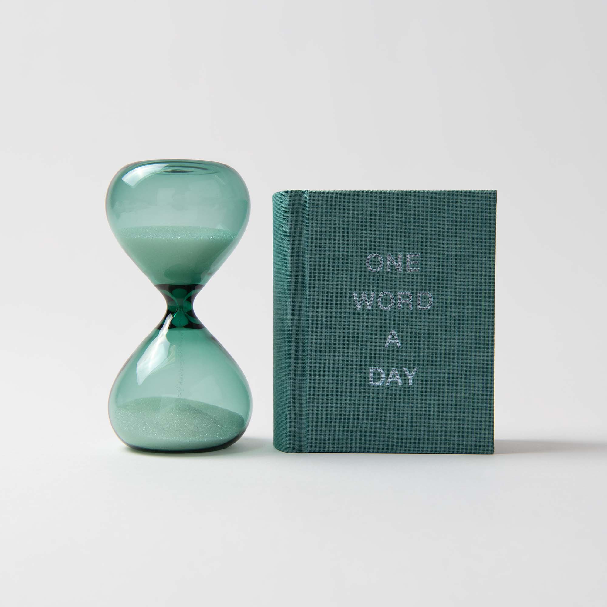 Das handgefertigte „One Word a Day mit Sanduhr“ von paperlux.store zeigt neben dem Buch eine grüne, mit Sand gefüllte Sanduhr auf weißem Hintergrund – und ist damit ein ideales, einzigartiges Geschenk für jeden Anlass.