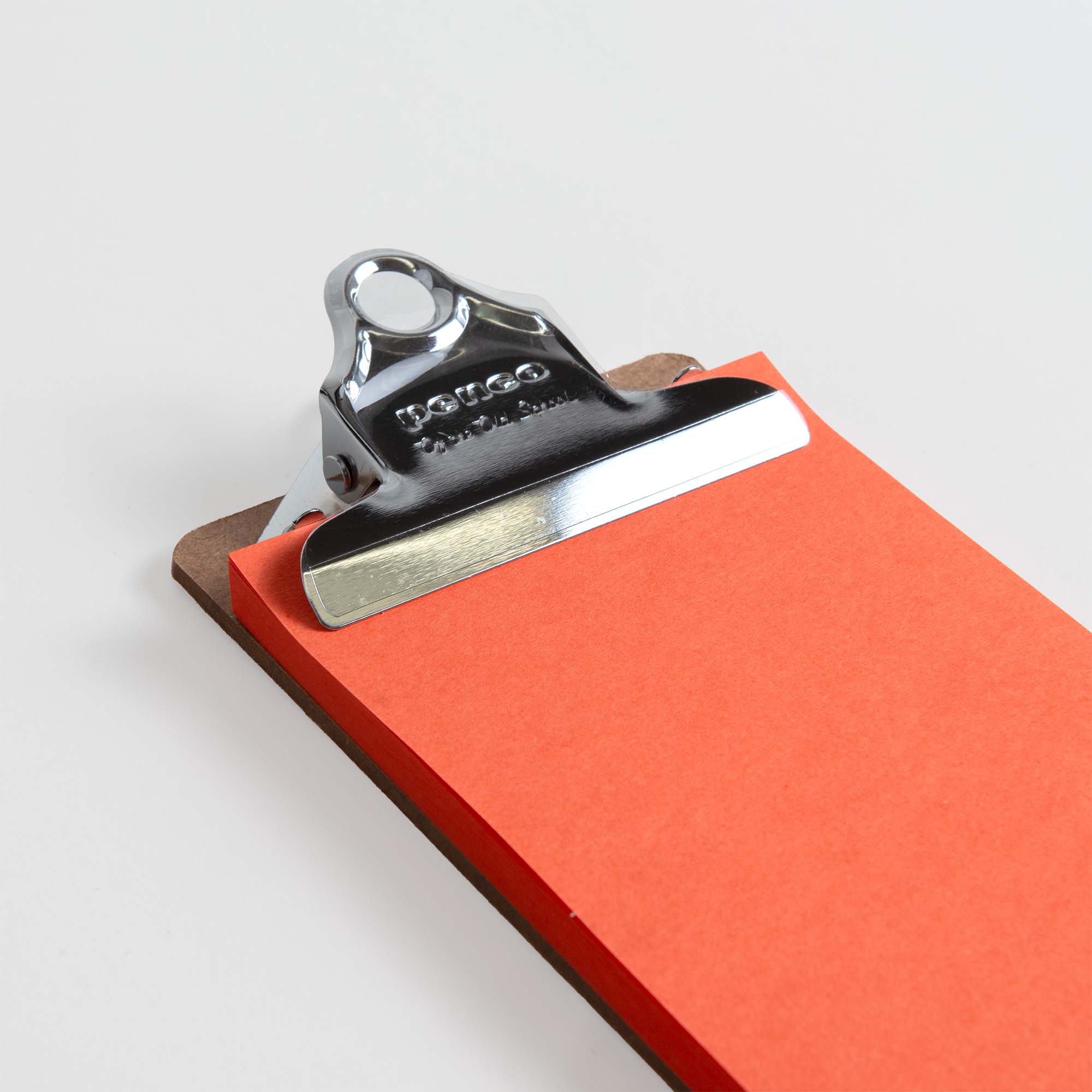 Eine Nahaufnahme des Penco Clipboard XS - Silber von Penco mit einer Metallklammer, die leuchtend orangefarbene Papierblätter auf einer weißen Oberfläche hält.