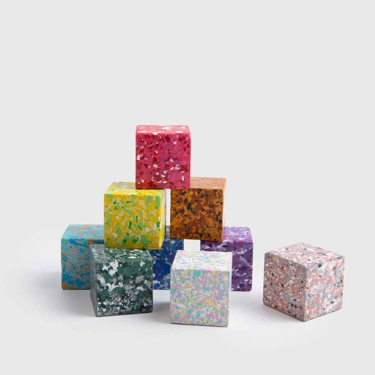 Acht lebhafte, gesprenkelte „Color Gem – das Wachsmalquadrat“-Würfel von Studio Unto sind zu einer Pyramide angeordnet, wobei ein weiterer Würfel rechts davon platziert ist. Inspiriert von „Zeichnen aus der Natur“ weist jeder Würfel eine einzigartige Kombination aus leuchtenden Farben und Mustern auf, die sich alle vor einem schlichten hellgrauen Hintergrund abheben.
