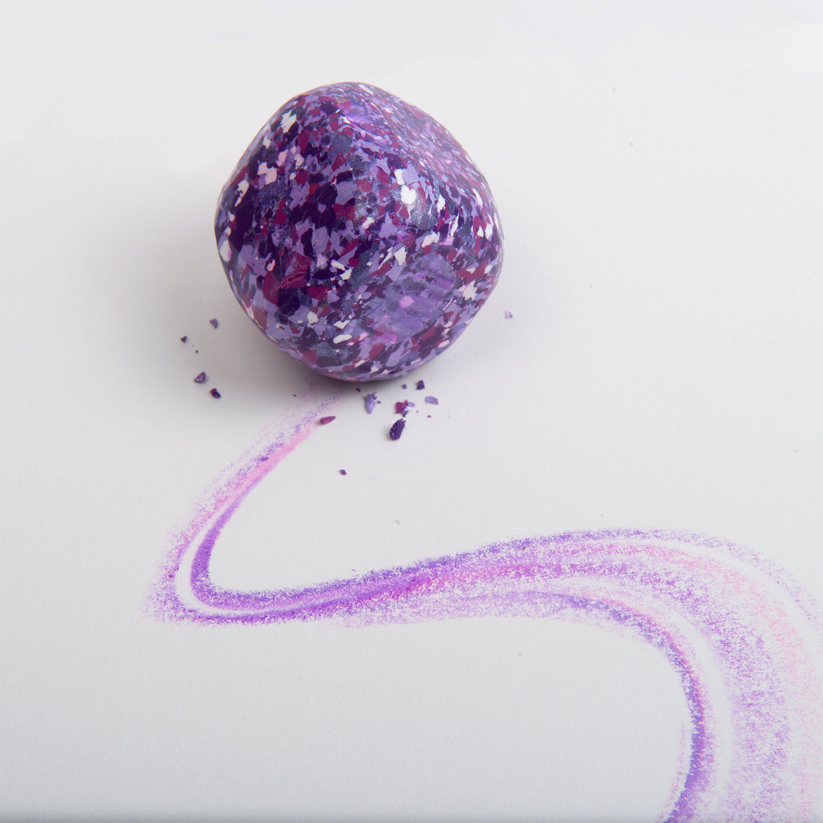 Ein leuchtendes, glitzerndes lila Wachsmalquadrat von Studio Unto – Color Gem – das Wachsmalquadrat Einzelpackung – hat eine wellenförmige, lila Linie auf weißem Papier hinterlassen. Zarte lila Flocken sind daneben verstreut, was an das „Zeichnen aus der Natur“ erinnert und jeden dazu inspiriert, Kunstwerke voller farbenfroher Kreativität zu schaffen.