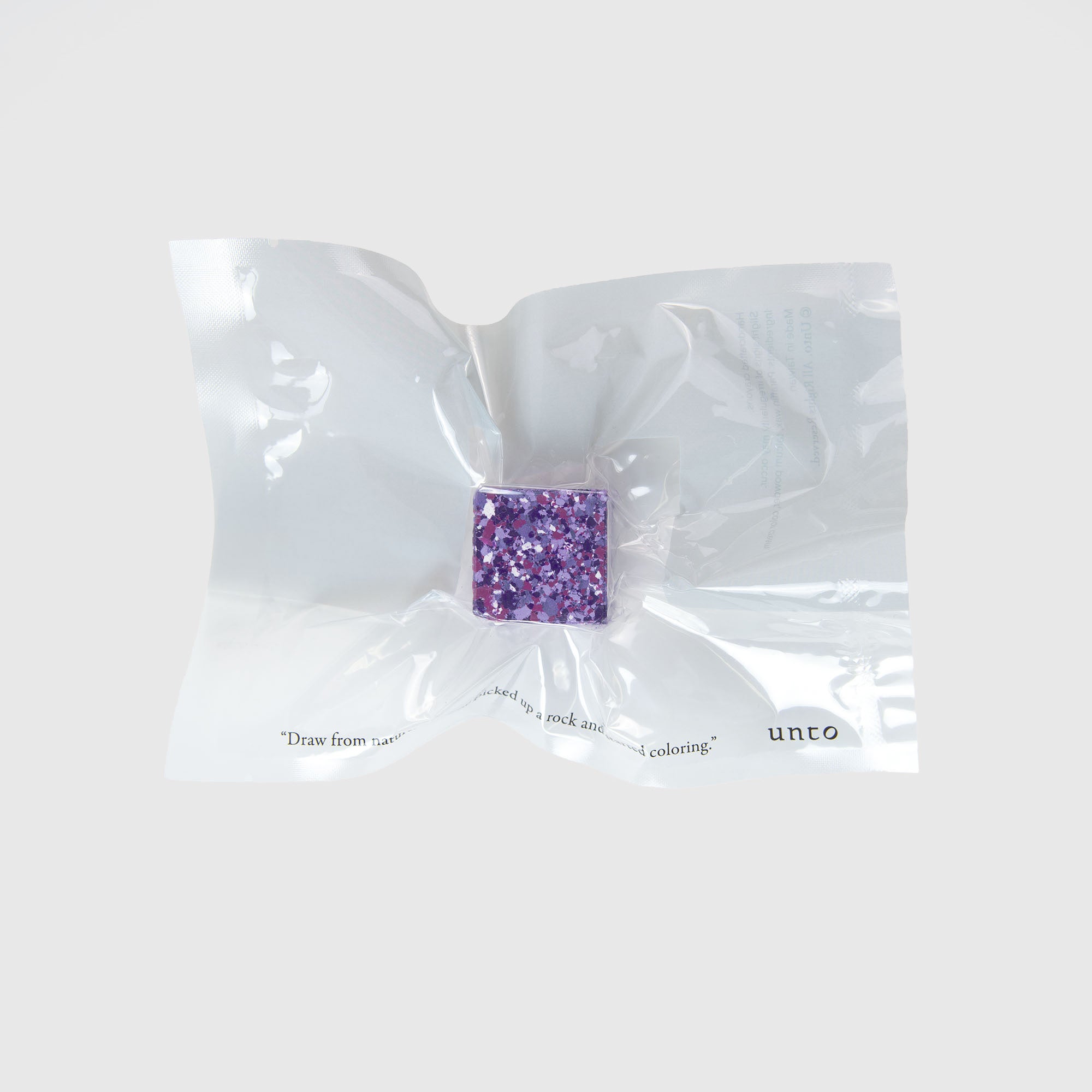 Eine vakuumversiegelte Packung enthält einen kleinen quadratischen Color Gem von Studio Unto mit einem violetten, gesprenkelten Muster. Die durchsichtige Verpackung ist leicht zerknittert und auf ihr ist ein Text auf einem hellgrauen Hintergrund zu sehen.