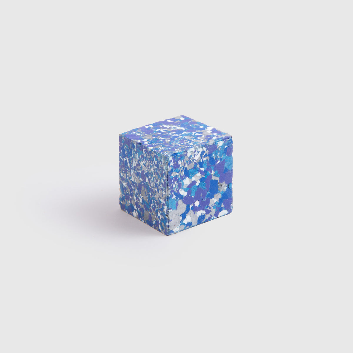 Ein kleines, blau gesprenkeltes „Color Gem“ – das Wachsmalquadrat – Einzelpackung von Studio Unto, dessen Oberfläche mit verschiedenen Blau- und Weißtönen übersät ist, ruht auf einem schlichten hellgrauen Hintergrund. Seine filigranen Muster fangen die natürliche Kunstfertigkeit ein, die das Zeichnen aus der Natur hervorruft.