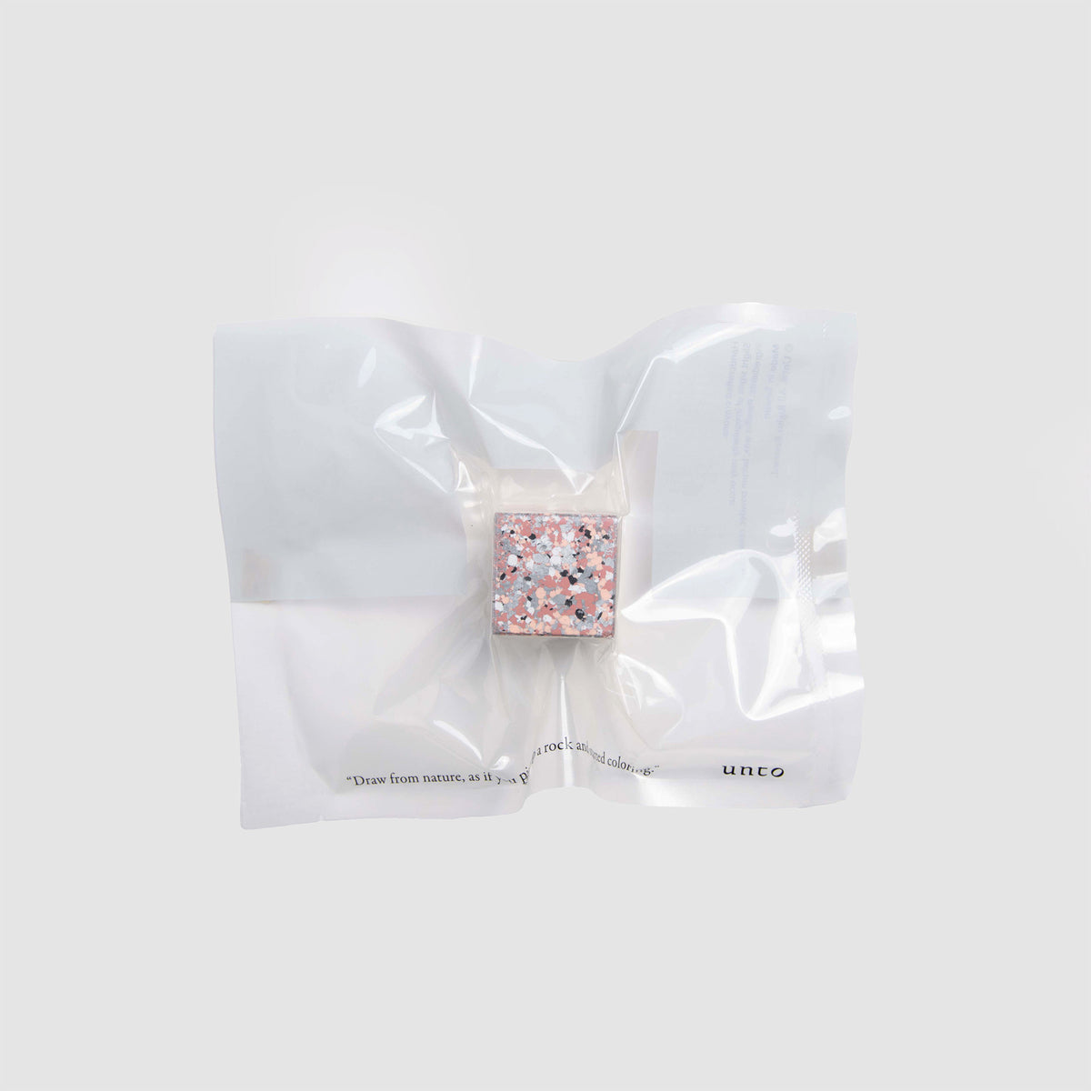 Ein kleines, quadratisches, mehrfarbiges Terrazzo-Objekt – „Color Gem – das Wachsmalquadrat“ Einzelpackung von Studio Unto – ist in einem durchsichtigen, vakuumversiegelten Plastikbeutel verpackt und vor einem schlichten hellgrauen Hintergrund platziert.