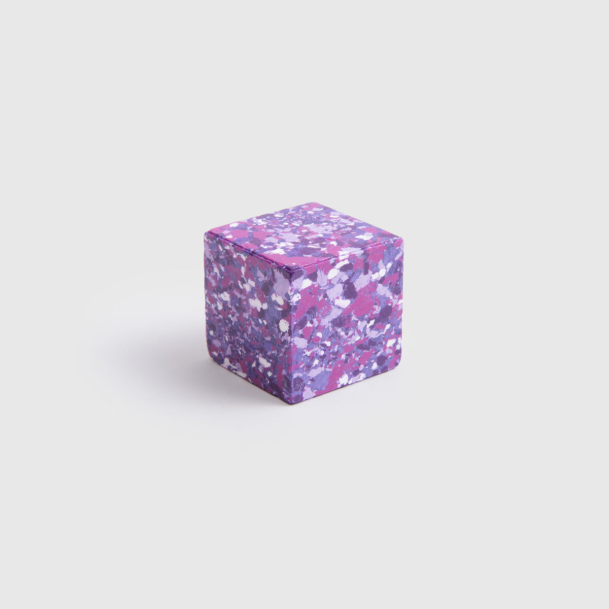 Ein kompaktes, violettes „Color Gem“ – das „Wachsmalquadrat“ von Studio Unto mit einem gesprenkelten Muster aus weißen, rosa und dunkleren violetten Flecken – ruht auf einem schlichten hellgrauen Hintergrund und erinnert an ein einzigartiges künstlerisches Werkzeug, das zum Zeichnen aus der Natur oder zum Erstellen von Kunstwerken inspiriert.