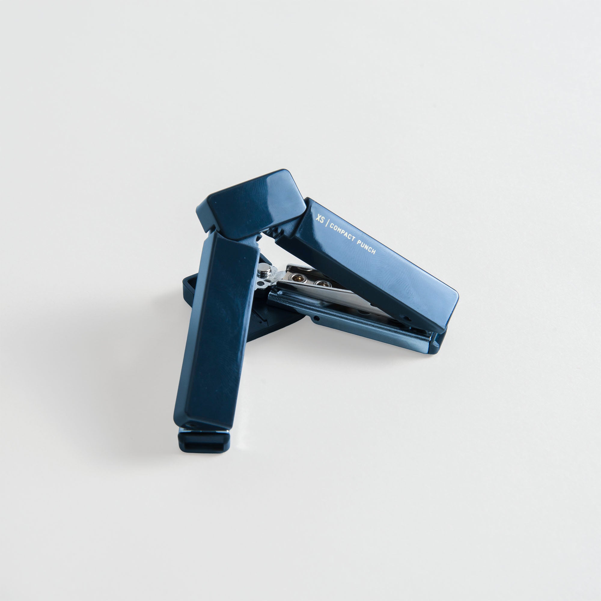 Auf einer weißen Fläche liegt schräg ein dunkelblauer Hefter mit sichtbaren Metallteilen, daneben der „Midori XS Compact Punch“ von Midori – der kleinste Locher mit integriertem Chiphalter.