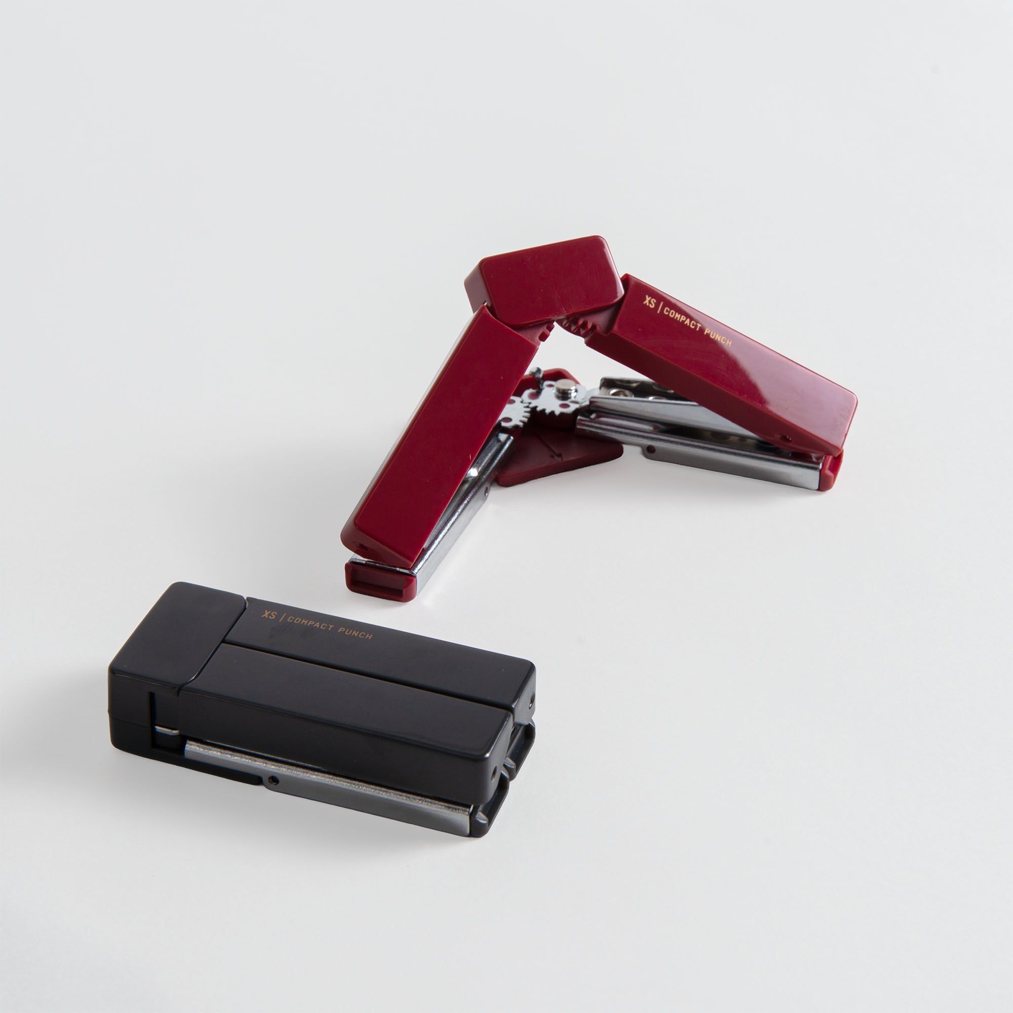 Zwei kompakte Hefter, ein schwarzer und ein weinroter, stehen auf einer weißen Fläche. Der weinrote Hefter ist teilweise geöffnet und offenbart seinen inneren Mechanismus - ideal für die Verwendung mit dem Midori XS Compact Punch von Midori.