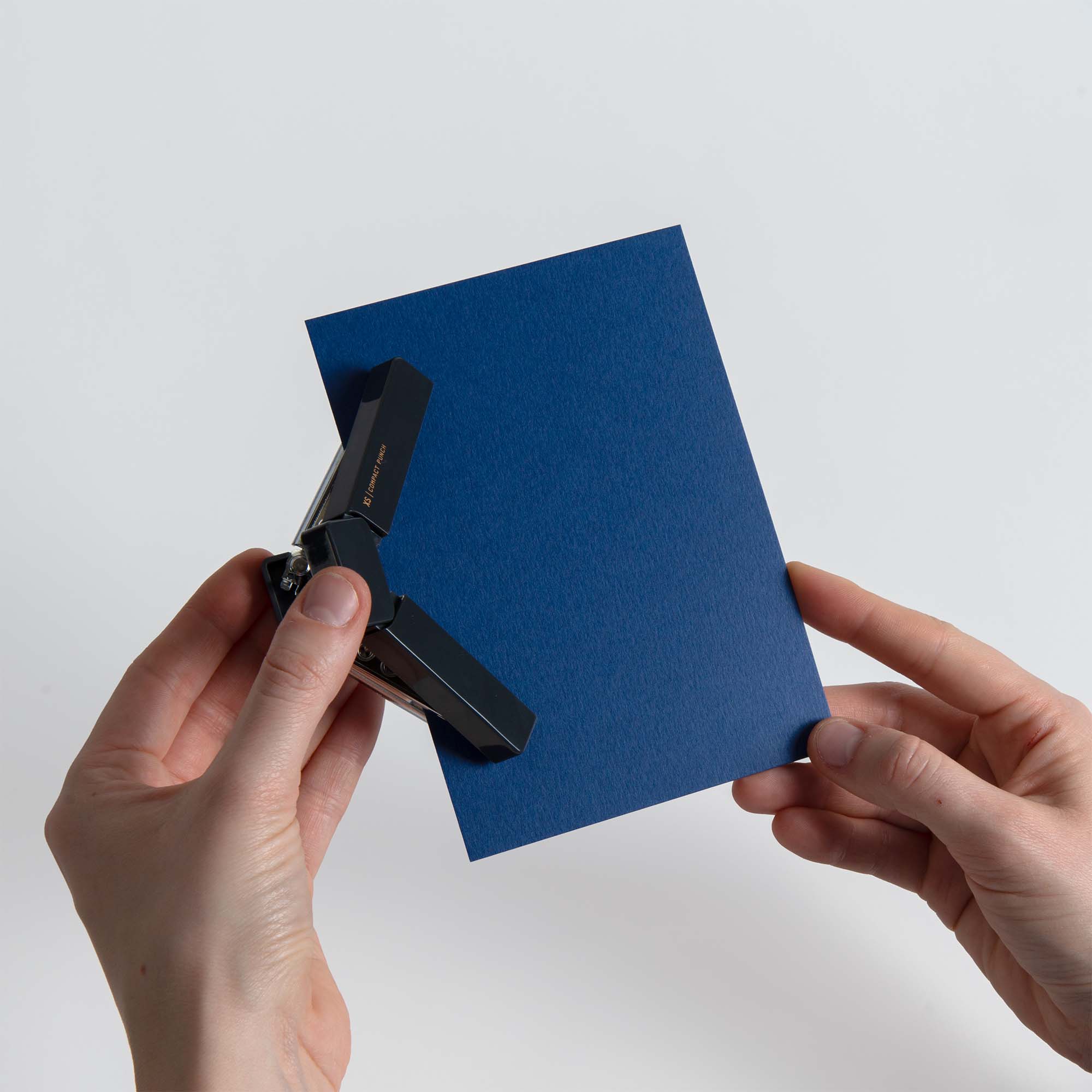 Eine Person hält eine blaue, rechteckige Karte in der Hand und stanzt mit dem Midori XS Compact Punch von Midori eine Ecke aus, wobei der Hintergrund einfarbig weiß ist.