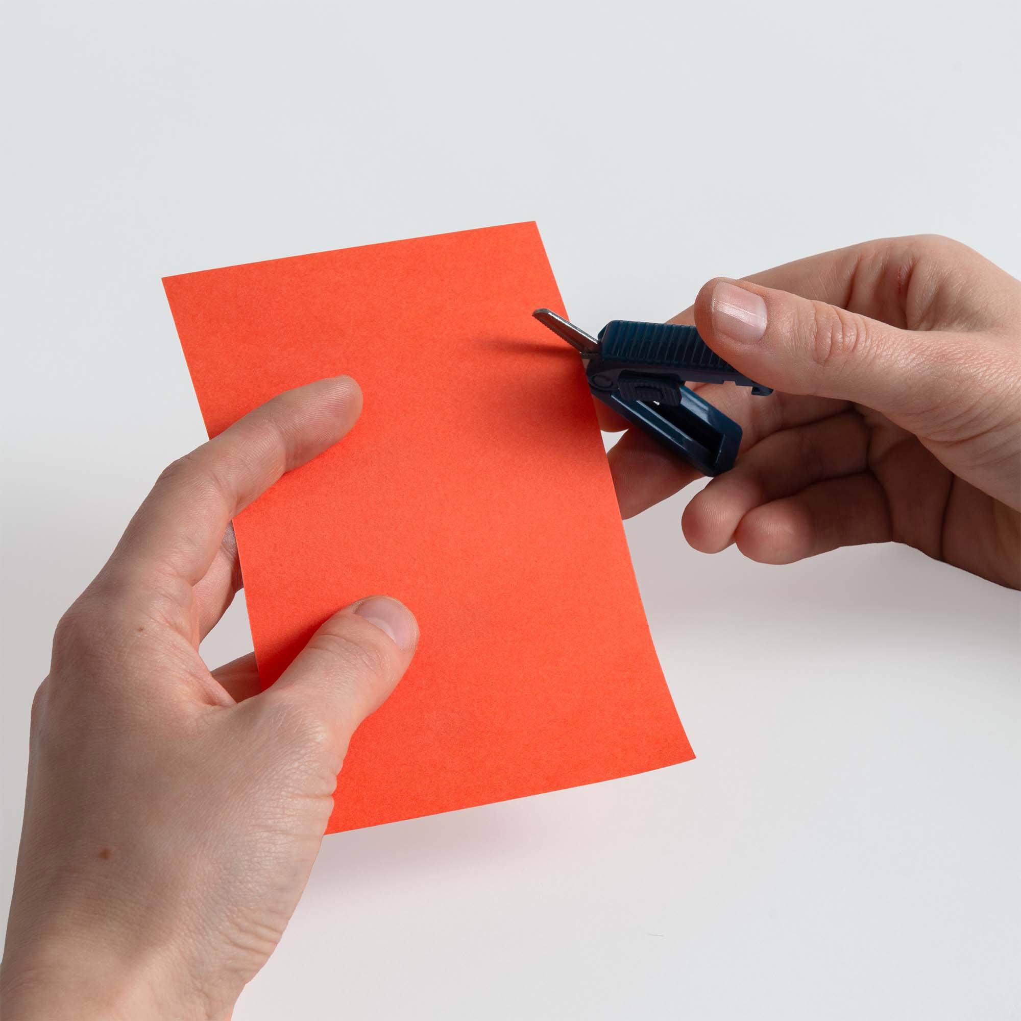 Eine Person hält ein Blatt rotes Papier in der einen Hand und eine schlanke Midori Kompaktschere mit Zacken in der anderen und bereitet sich darauf vor, das Papier vor einem schlichten weißen Hintergrund zu schneiden.