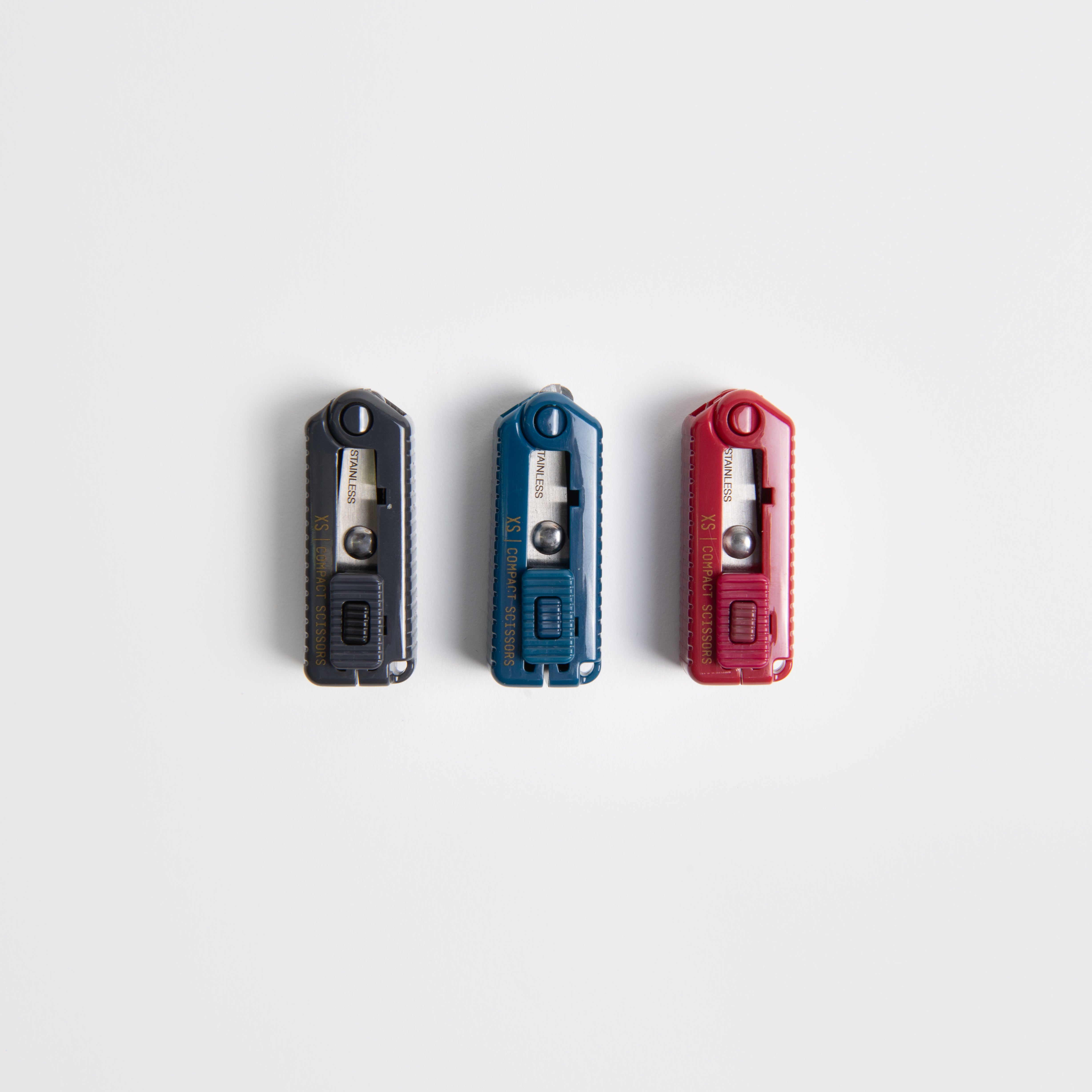 Drei kompakte Gebrauchsmesser - schwarz, blau und rot - sind vertikal auf einem weißen Hintergrund angeordnet. Jedes hat einen Schiebeknopf und eine freiliegende Klingenspitze, so tragbar wie die Midori Kompaktschere.