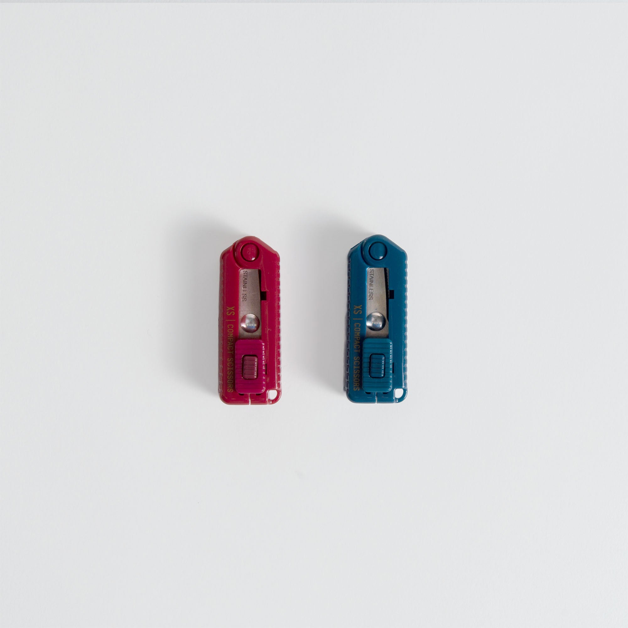 Zwei Compact Scissors Cerrated von Midori, eine rote und eine blaue, mit scharfen Klingen, sind nebeneinander auf einem schlichten weißen Hintergrund platziert.