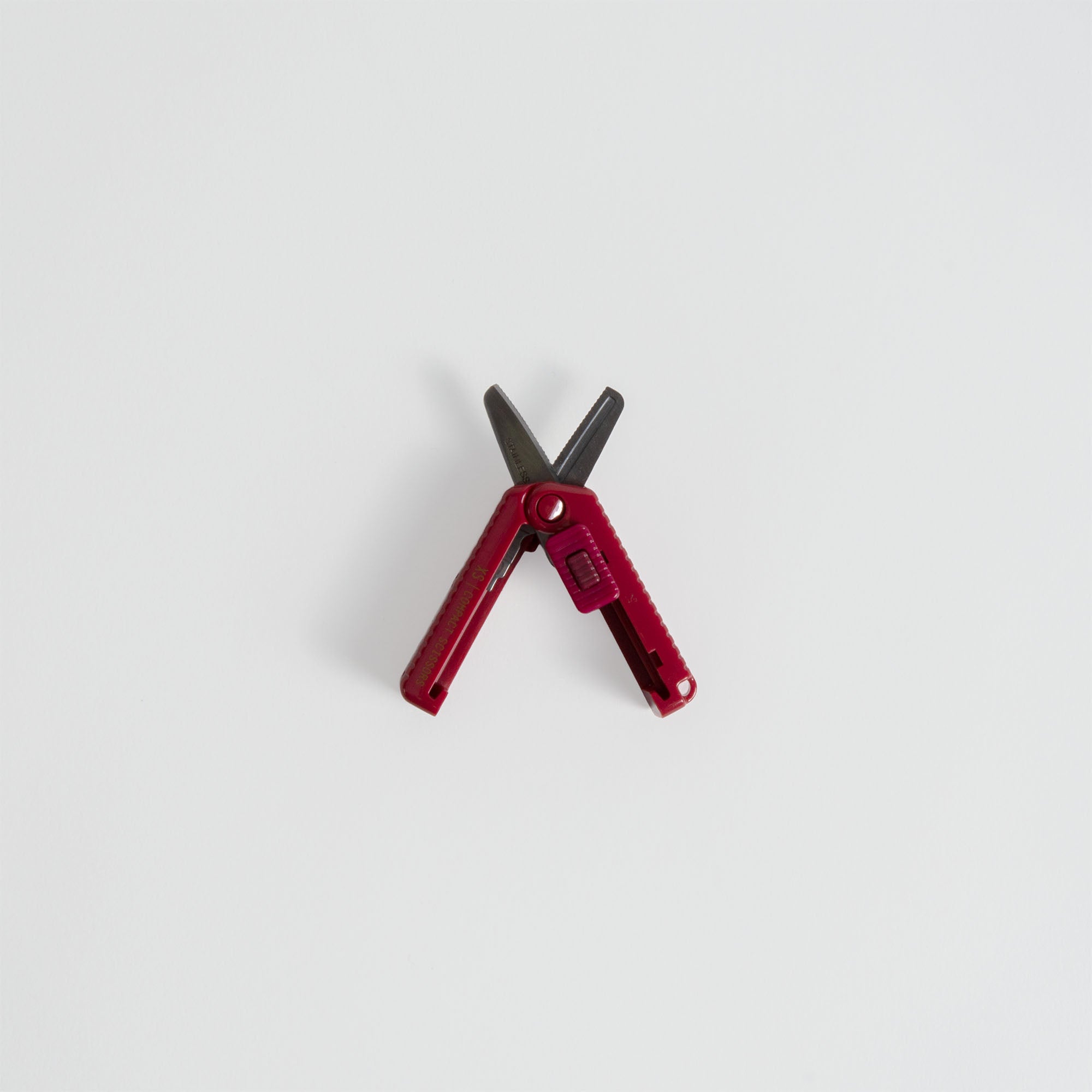 Eine Midori Kompaktschere mit roten Griffen und schwarzen Klingen liegt offen auf einer schlichten weißen Fläche.
