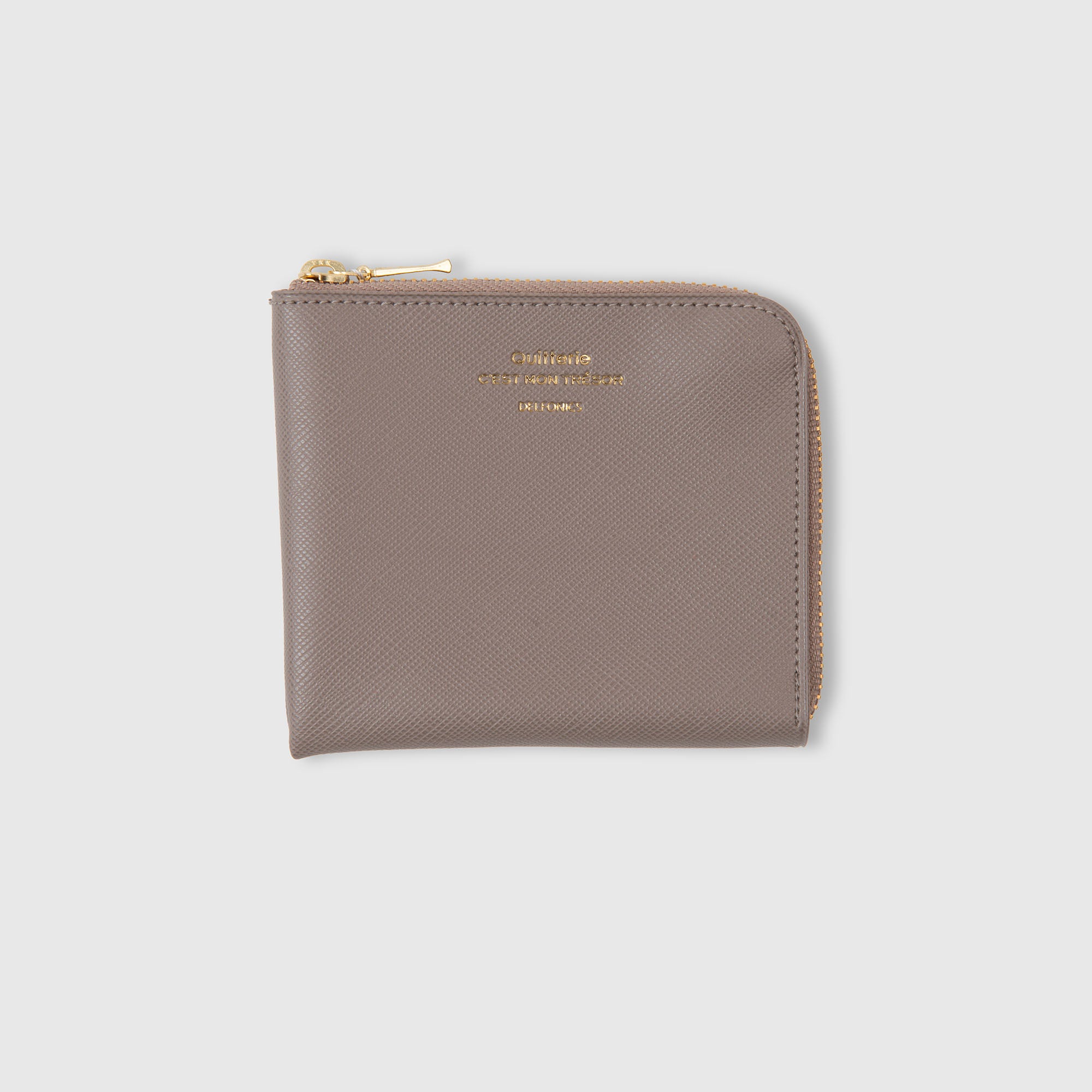 Ein taupefarbenes Delfonics Quitterie Half Zip Card Case | Kartenetui aus Leder im Scotch-Grain-Look mit goldenem Reißverschluss und geprägtem goldenem Schriftzug auf der Vorderseite, der sich von einem schlichten hellgrauen Hintergrund abhebt. Dieses stilvolle Portemonnaie ist zudem wasserabweisend und somit besonders strapazierfähig.