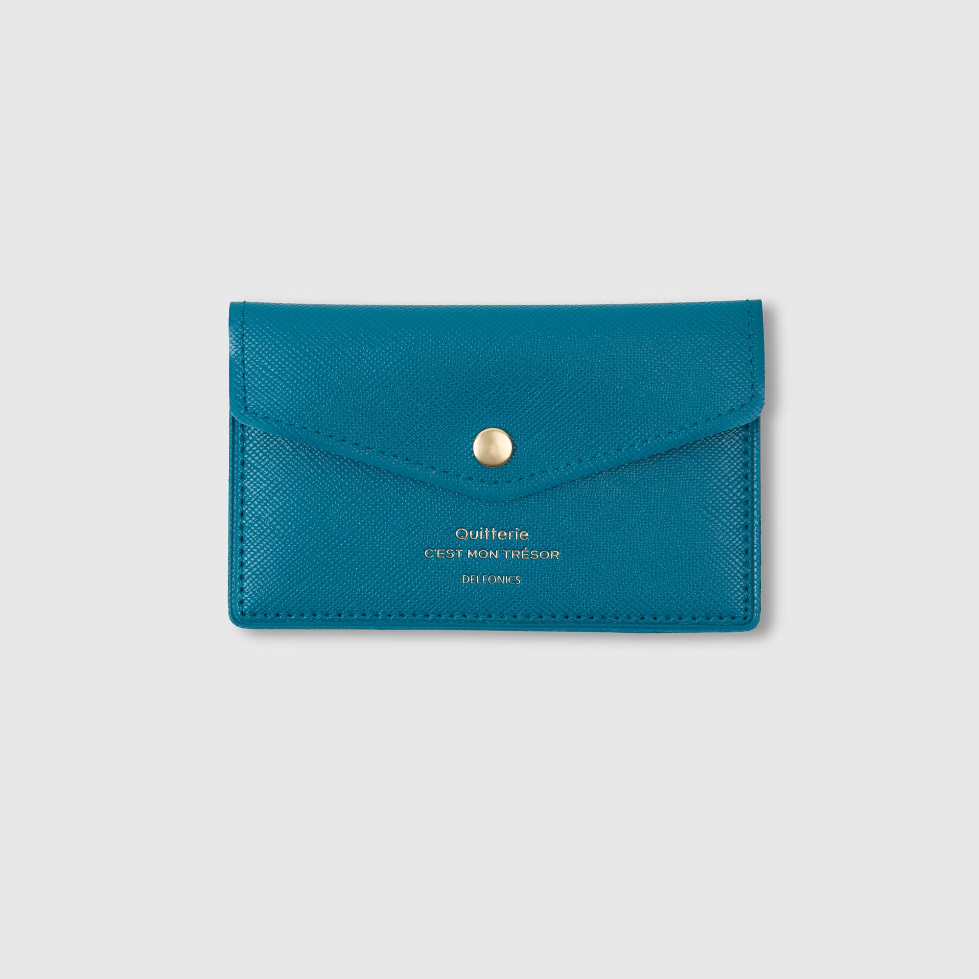 Eine türkisfarbene Lederbrieftasche im Umschlagstil von Delfonics mit dem Namen „Quitterie Card Case“ Kartenetui verfügt über einen goldenen Druckknopf und eine elegante goldene Aufschrift – „Quiltérie CEST MON TRESOR DELFINES“ – auf der Vorderseite, die sich von einem schlichten hellgrauen Hintergrund abhebt.