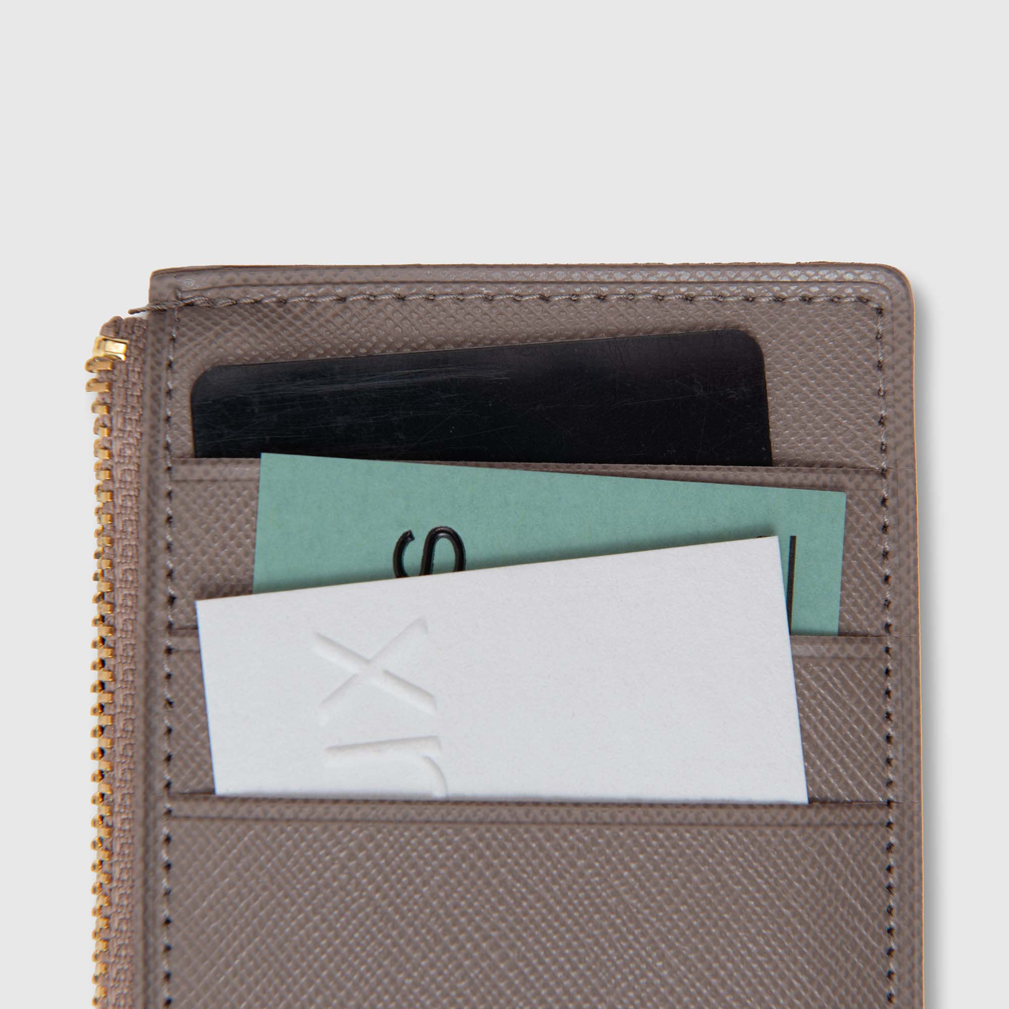 Eine Nahaufnahme eines grauen „Delfonics Quitterie Card Zip Case | Kartenetui“, in dessen Kartenfächer neben dem Reißverschluss drei Karten – eine schwarze, eine grüne und eine weiße – teilweise eingesteckt sind.