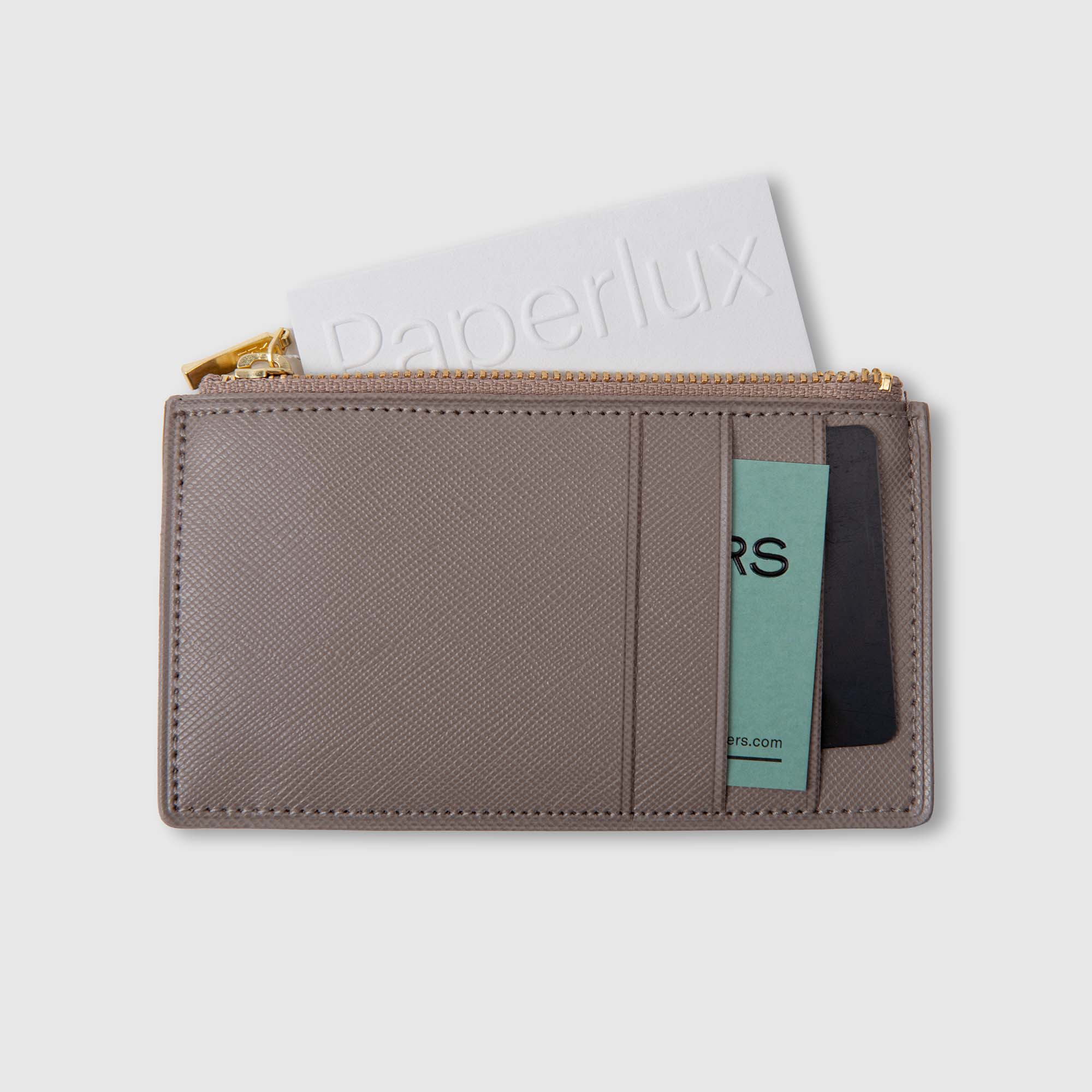 Ein taupefarbenes Delfonics Quitterie Card Zip Case | Kartenetui aus Leder von Delfonics verfügt über einen goldenen Reißverschluss und bietet Platz für eine weiße Karte mit der Aufschrift „Paperlux“ sowie zwei weitere Karten – eine grüne und eine schwarze – in den Kartenfächern.