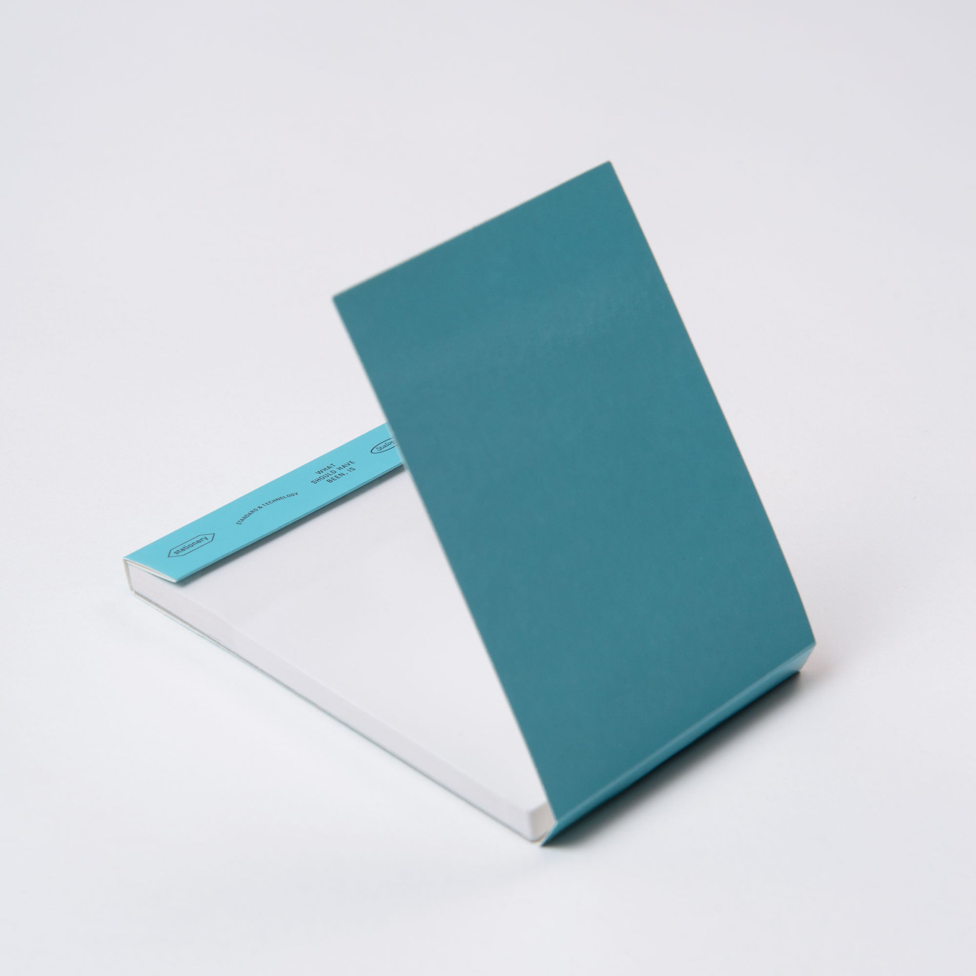 Ein blaugrüner Editors Memo Pad von Stalogy liegt auf einer weißen Oberfläche. Sein Einband ist teilweise geöffnet und gibt den Blick auf eine leere weiße Seite frei. Die scharfen Kanten und die leichte Neigung verleihen ihm eine gelassene Eleganz.