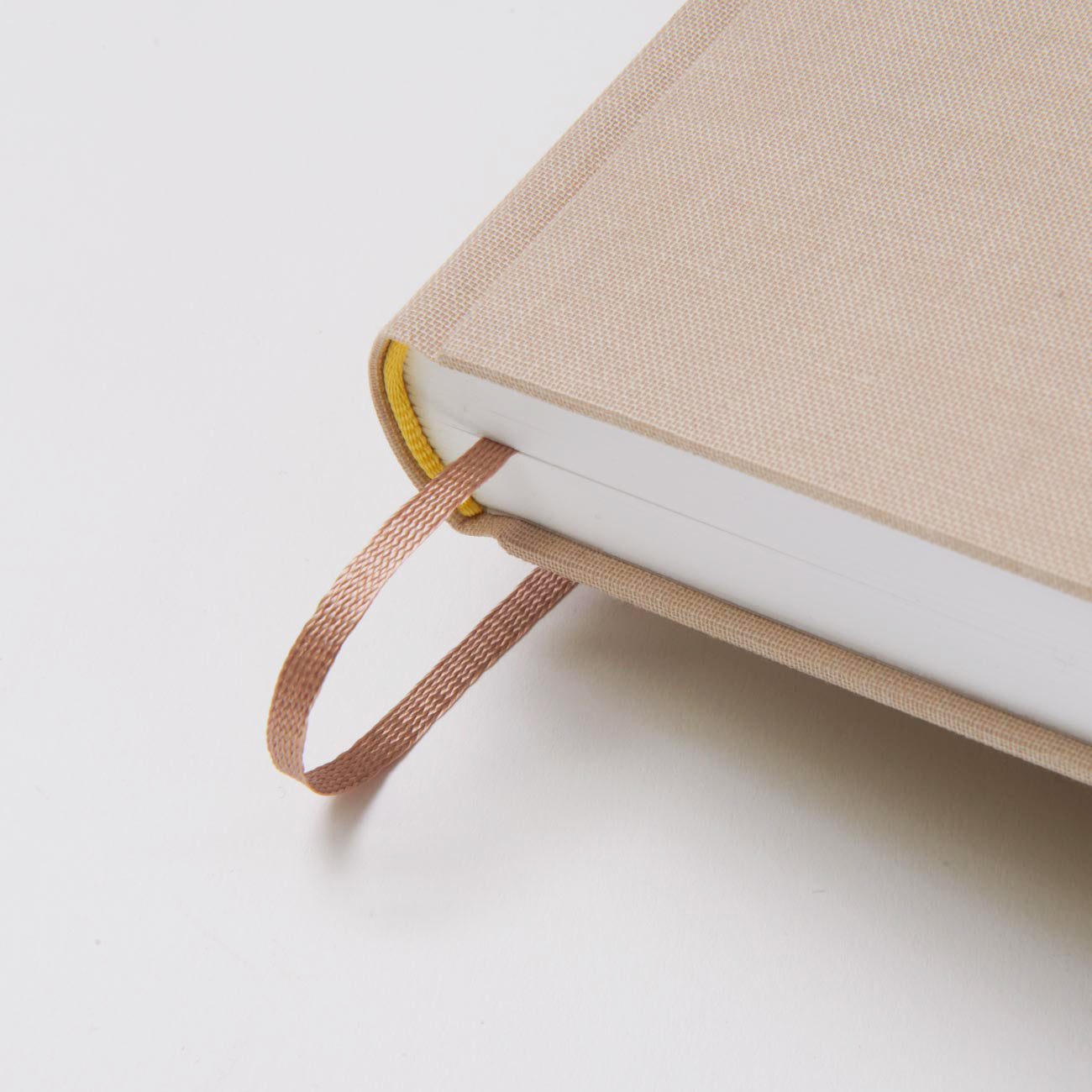 Nahaufnahme der Ecke des beigen Hardcover-Buchs „Gästebuch – Willkommen in unserer Krippe“ von We are Bookbinders, Made in Hamburg, auf weißem Untergrund. Ein braunes Lesebändchen ragt aus der oberen Ecke hervor, die Textur des Buchdeckels ist sichtbar, der Buchrücken ist gelb akzentuiert.