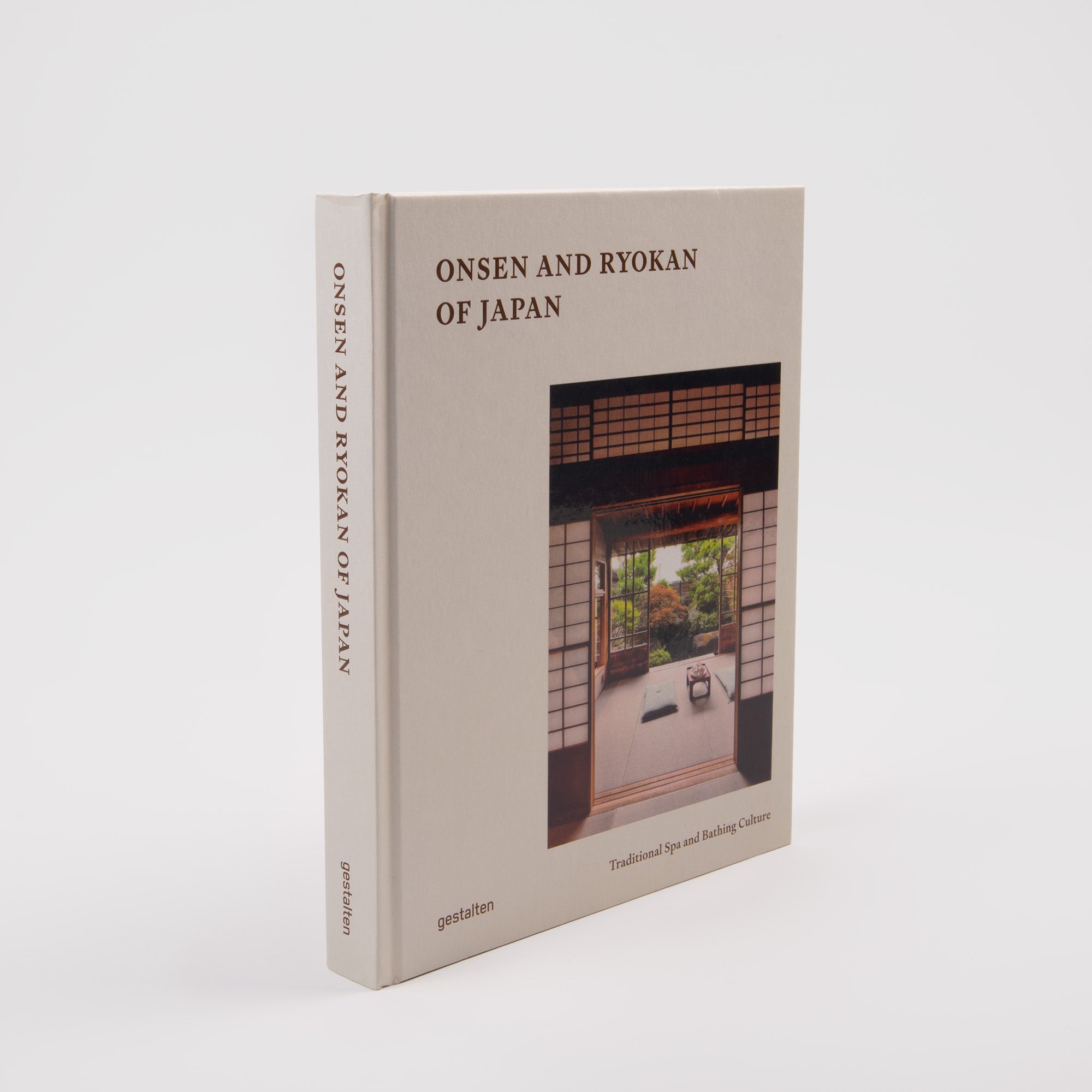 Ein beigefarbener Hardcover-Einband mit dem Titel "Onsen & Ryokan of Japan - Ein Blick in Japans stille Welten" aus dem Gestalten Verlag steht aufrecht und zeigt ein traditionelles japanisches Zimmer mit Blick auf einen Garten - eine echte japanische Gastfreundschaft.