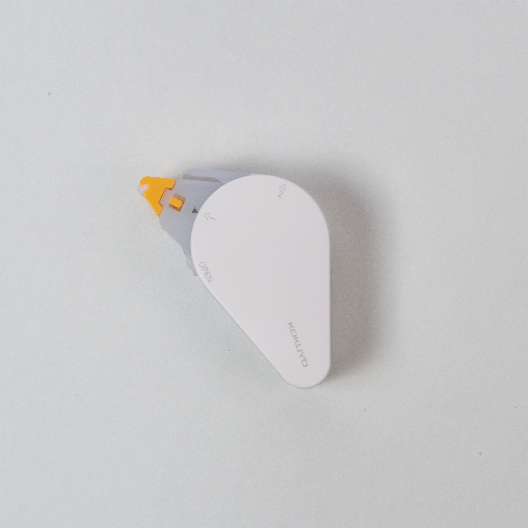 Der Kokuyo GLOO Tape Klebestreifen repositionierbar, danach permanent ist ein kleiner, eleganter weiß-grauer Spender auf hellgrauem Hintergrund. Seine Tropfenform und die orangefarbene Spitze unterstreichen seine Eleganz beim Abrollen des Klebebands.