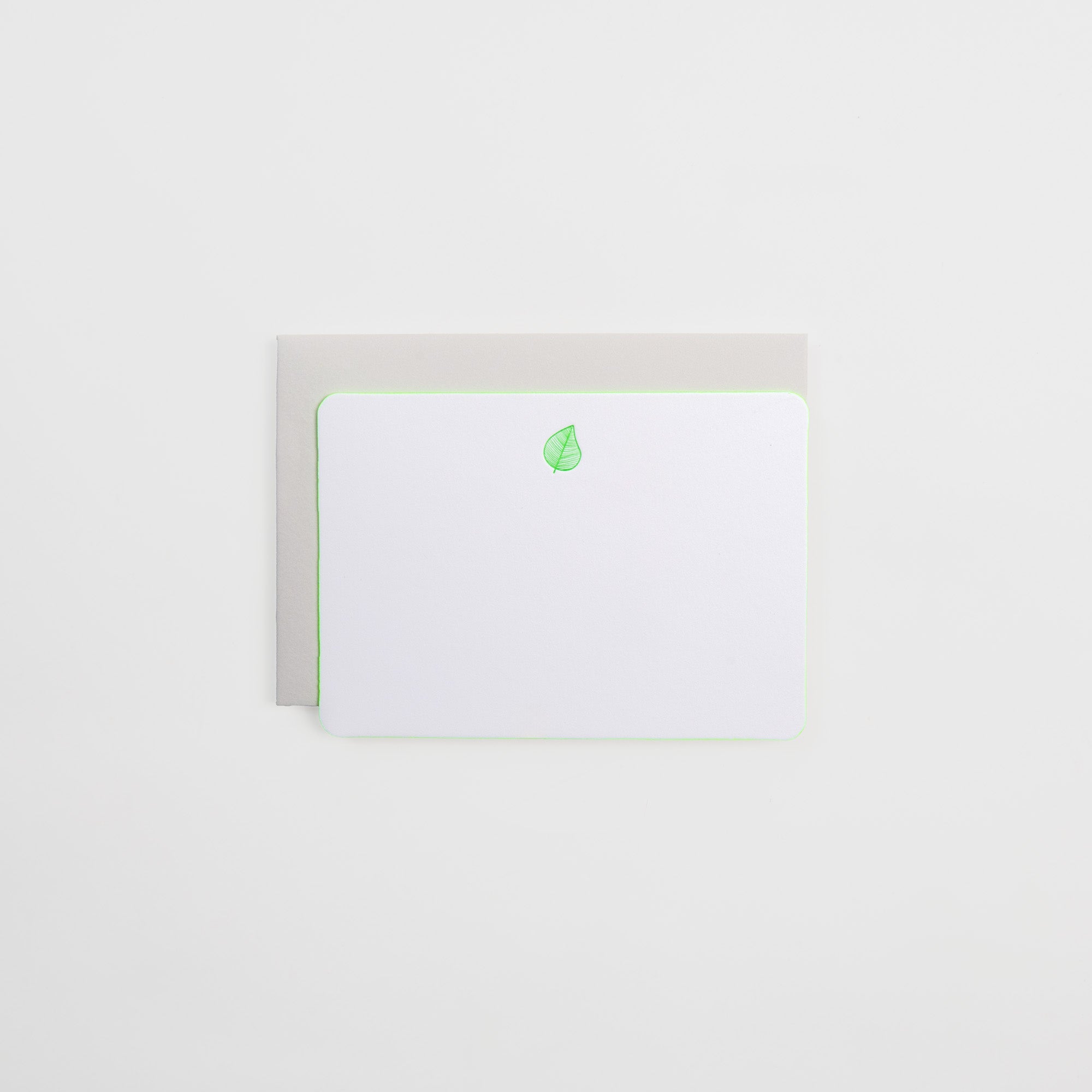 Die Grußkarte Icon mit Kuvert von Gmund ist eine weiße Blanko-Notizkarte aus Naturpapier mit abgerundeten Ecken und einem kleinen grünen Blattsymbol am oberen Rand, das auf einem hellgrauen Umschlag vor einem schlichten weißen Hintergrund ruht.