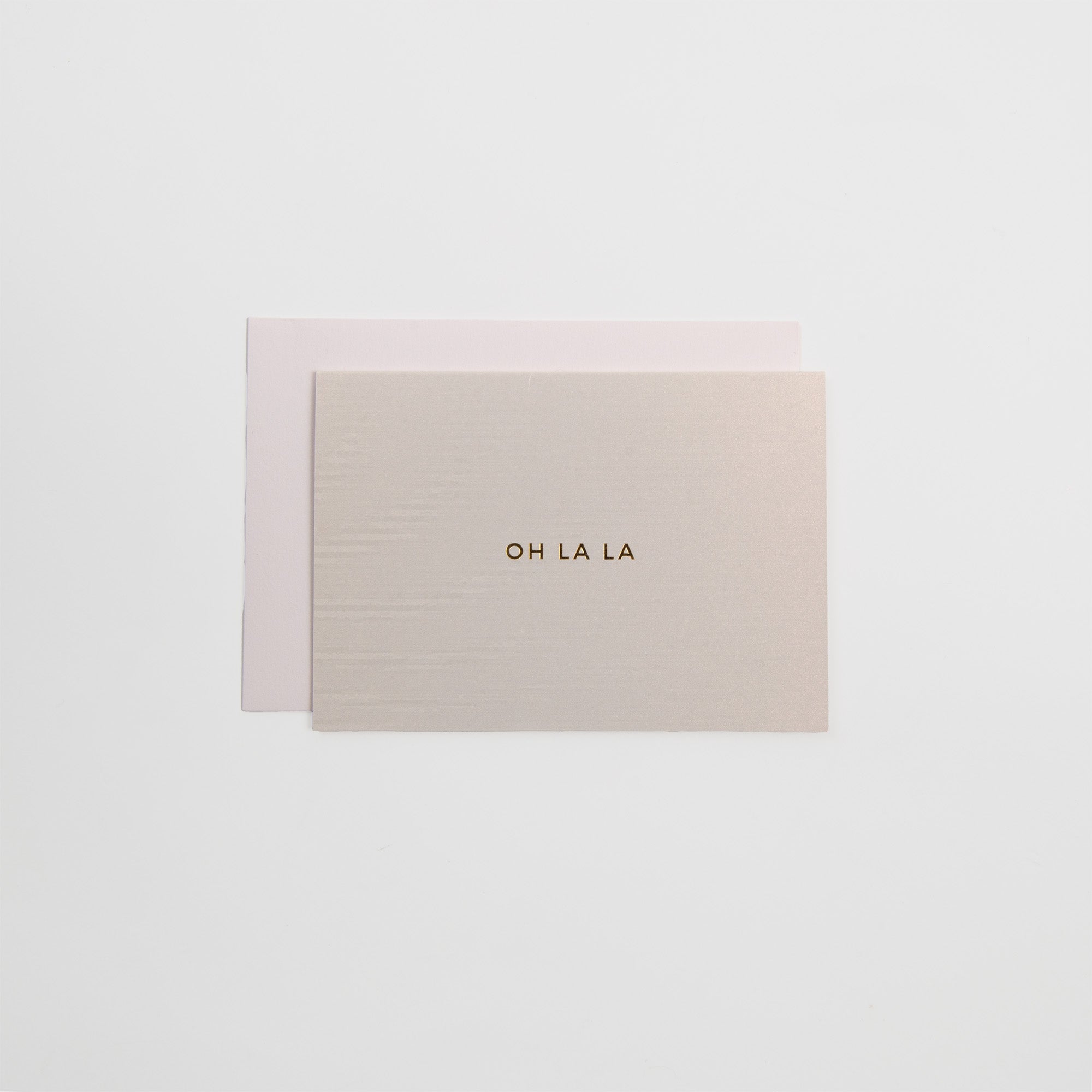 Das Kärtchen OH LA LA von Gmund zeigt einen goldenen Letterpress-Text auf beigem A7-Karton, gepaart mit einem hellrosa Gmund-Umschlag, der auf weißem Hintergrund steht.