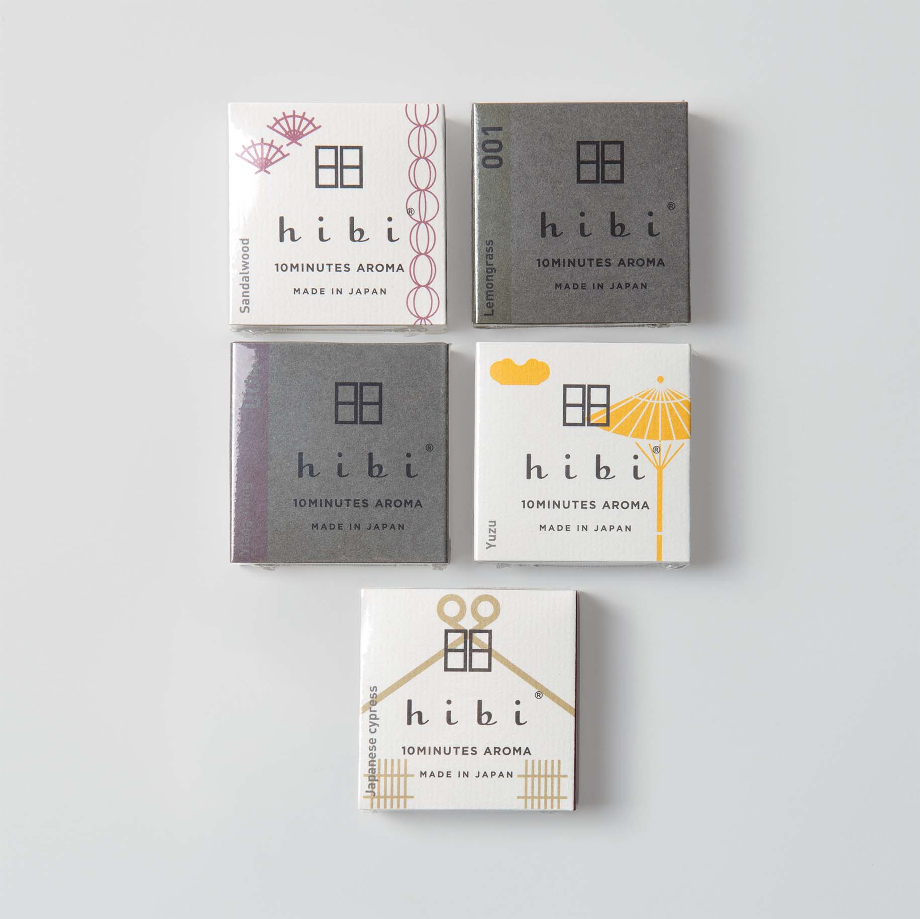 Fünf Schachteln Hibi - Duftstreichhölzer, die an japanische Streichhölzer erinnern, sind auf einer weißen Fläche angeordnet. Die minimalistische Verpackung zeigt einen Sonnenschirm und eine Schere mit Etiketten wie „001“, „Sandelholz“ und „Yuzu“. Auf den Schachteln steht „Made in Japan“, was ihren natürlichen Duft hervorhebt.