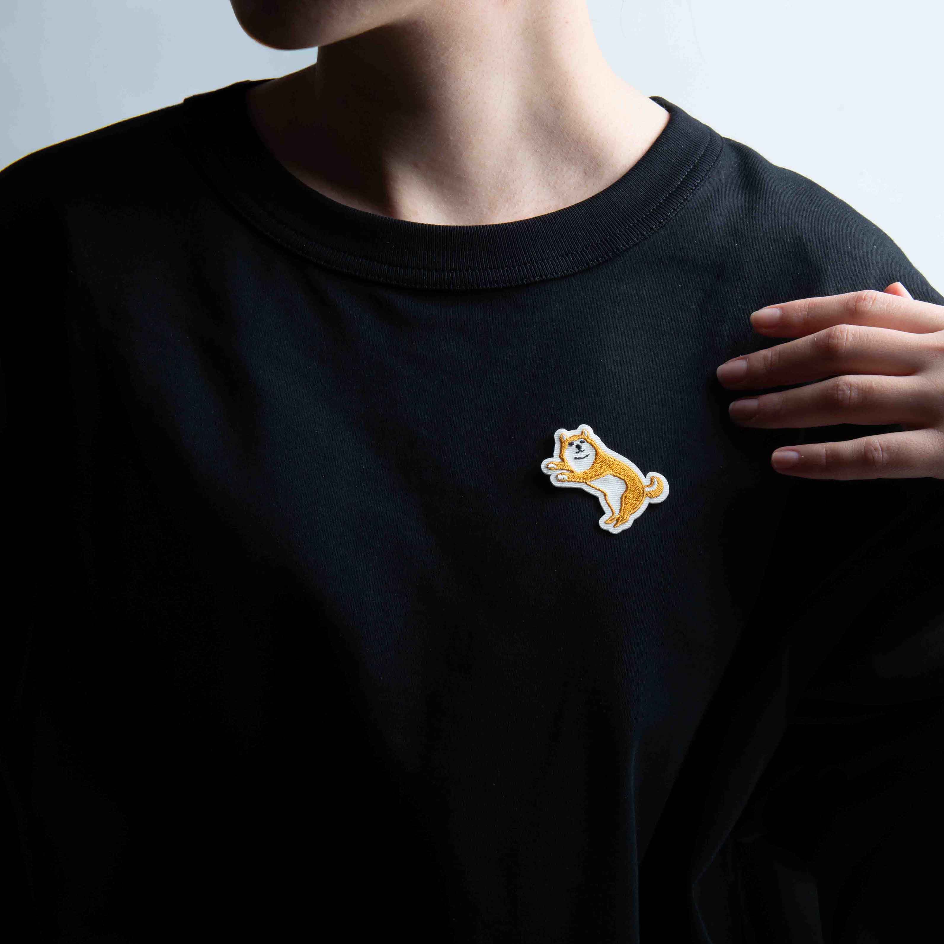 Eine Person in einem schlichten schwarzen Hemd trägt einen kleinen Hightide New Retro Patch Sticker - ein braunes und weißes Shiba Inu-Design von Hightide - auf der Brust, die Hand auf der Schulter, und versprüht den Charme der japanischen Showa-Ära.