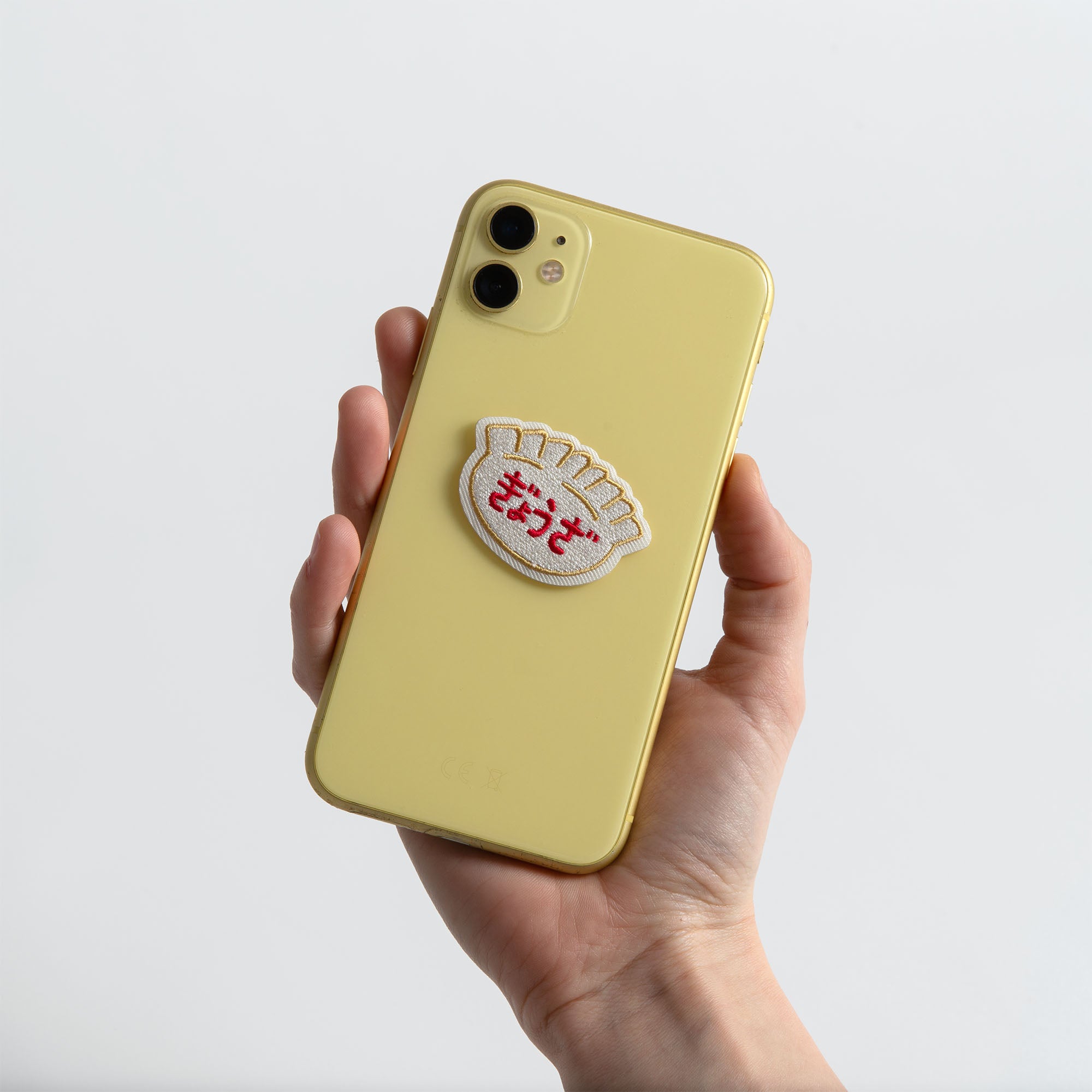 Eine Hand hält ein gelbes Smartphone, das mit dem Hightide New Retro Patch Sticker mit japanischem Text und einer verspielten Knödelform von Hightide verziert ist. Der schlichte weiße Hintergrund unterstreicht den nostalgischen Charme der Showa-Ära.