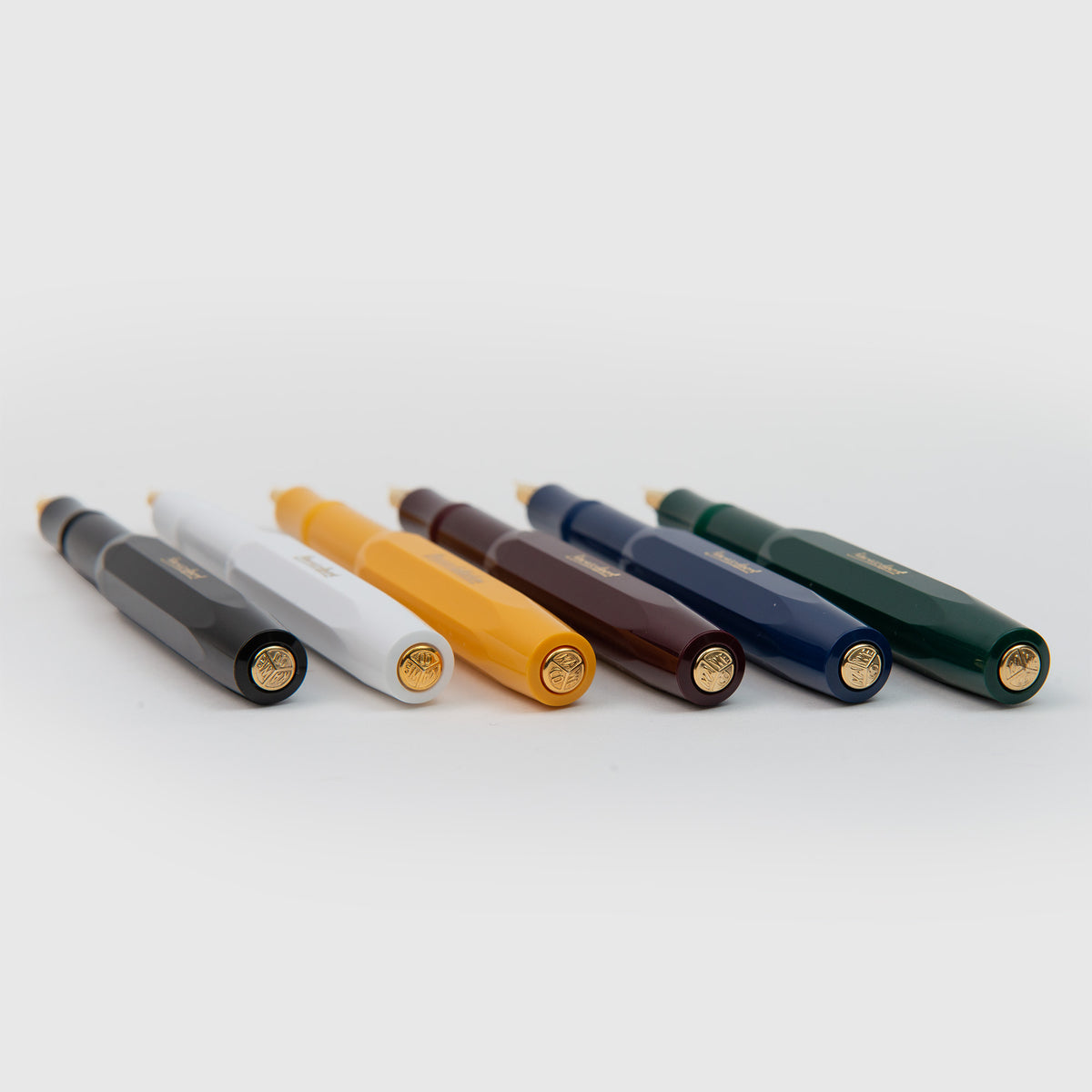 Sechs Kaweco Classic Sport – Füllhalter von Kaweco in Schwarz, Weiß, Gelb, Braun, Blau und Grün sind horizontal auf einem weißen Hintergrund aufgereiht, wobei ihre goldfarbenen Endkappen zur Kamera zeigen. Jeder Füllhalter zeichnet sich durch ein unverwechselbares Design und eine goldene Feder für ein geschmeidiges Schreibgefühl aus.