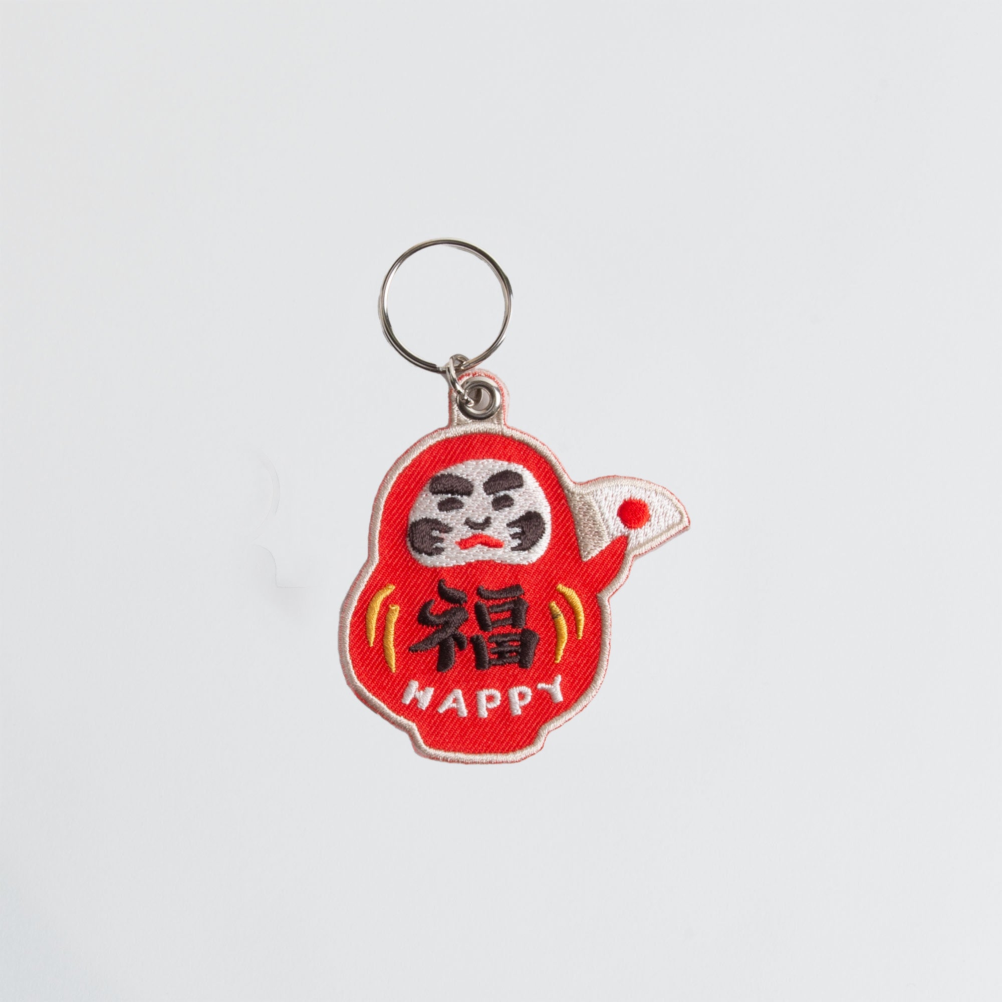 Der Hightide Stitch Work Keychain - Kamera, Daruma & Glückskatze von Hightide zeigt eine rote Daruma-Puppe mit gelben Akzenten und "HAPPY" in weiß - ein ideales Geschenk Japan, dargestellt auf einem schlichten weißen Hintergrund.