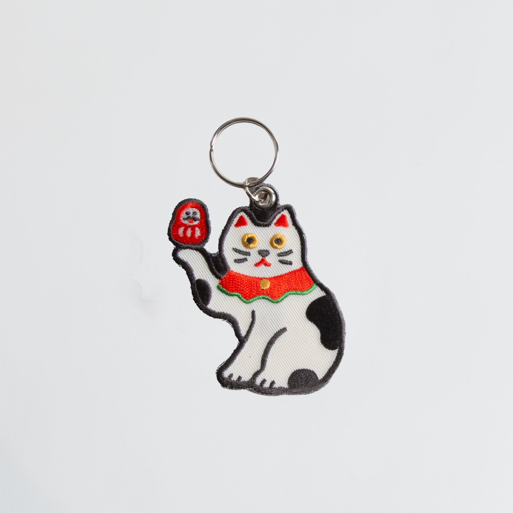 Der Hightide Stitch Work Keychain - Kamera, Daruma & Glückskatze von Hightide zeigt eine bestickte Glückskatze mit gelben Augen, rotem Halsband und roter Puppe - ideal als Geschenk für Japan-Fans.