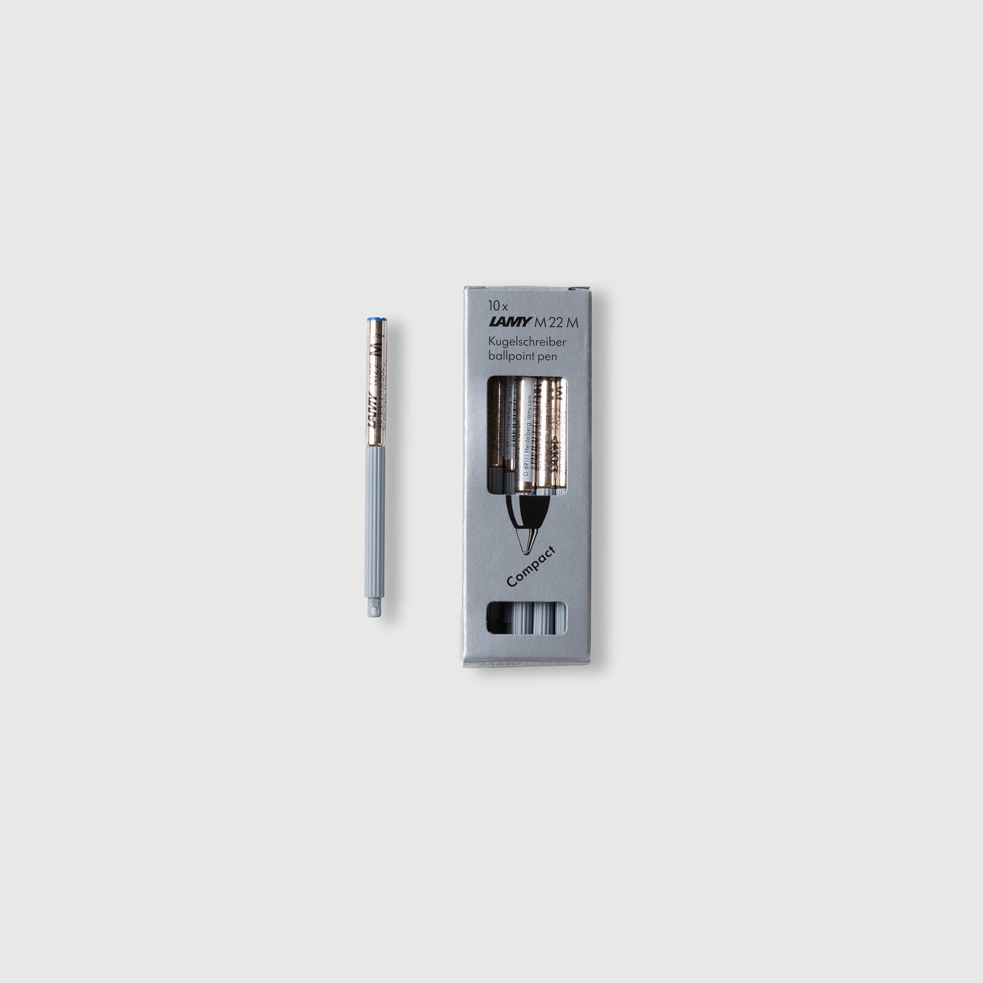 Ein silber-grauer Lamy pico Stift steht neben einer grauen Schachtel mit der Aufschrift "LAMY Compactmine M22 - für den LAMY pico" von Lamy, die mehrere Nachfüllpatronen enthält, alles auf einem schlichten weißen Hintergrund.
