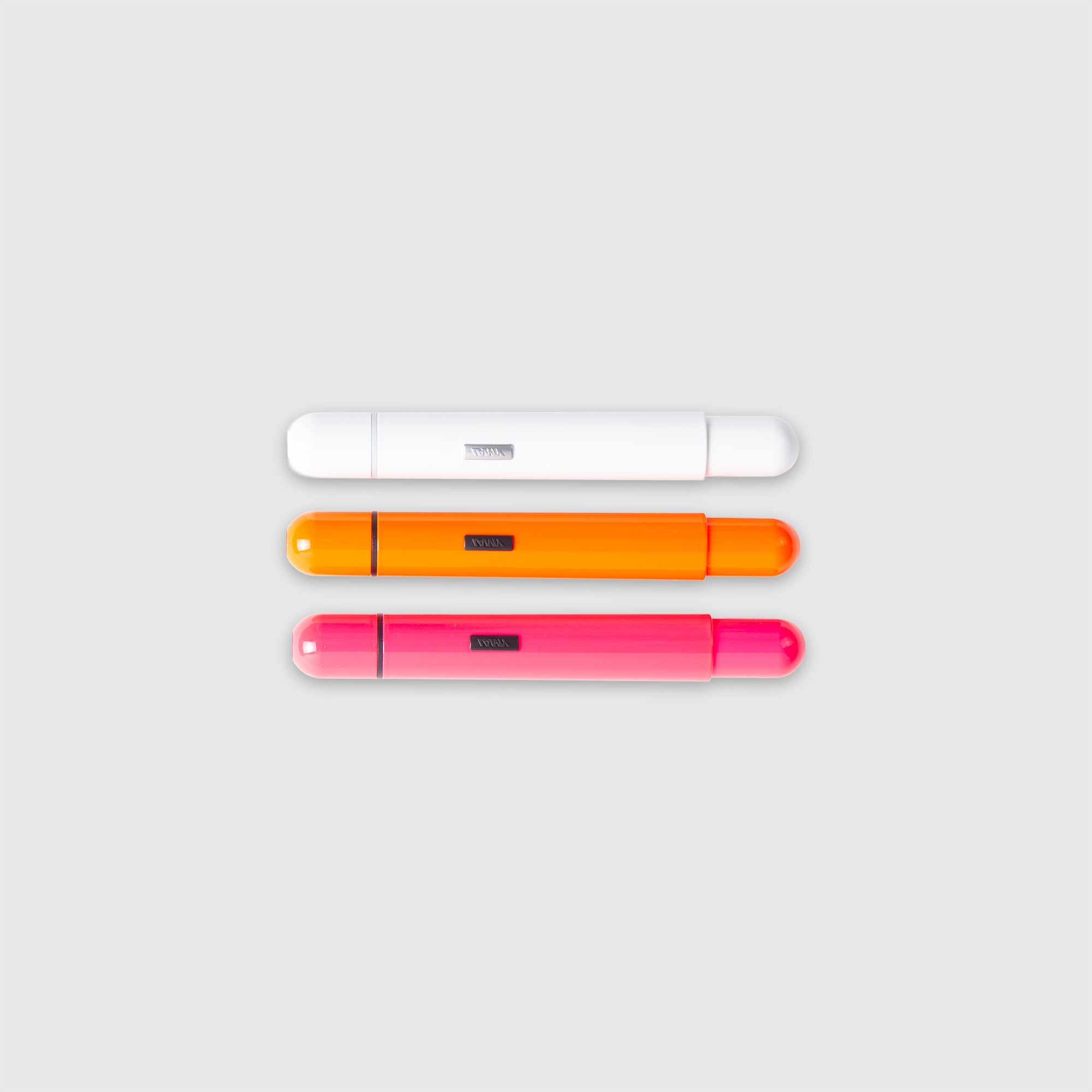 Drei kapselförmige LAMY pico Kugelschreiber von Lamy sind horizontal auf einem hellgrauen Hintergrund angeordnet. Die Stifte sind von oben nach unten weiß, orange und rosa und haben jeweils ein kleines dunkles Etikett in der Mitte.