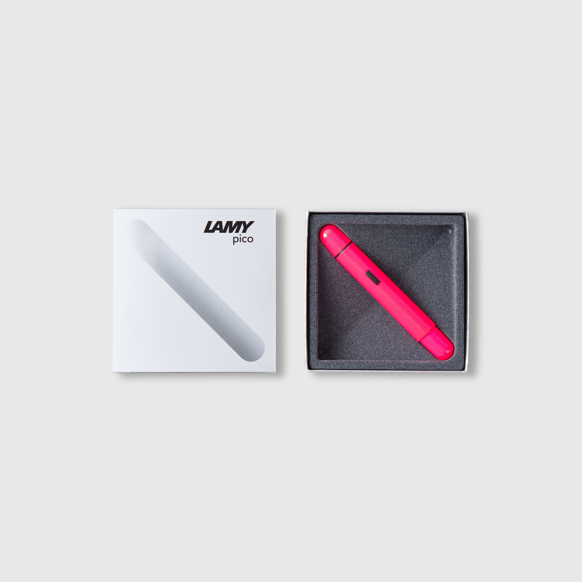Ein roter LAMY pico Kugelschreiber von Lamy wird in einer offenen grauen Schachtel neben seiner weißen Verpackung gezeigt, die ein diagonales transparentes Fenster und das Lamy-Logo auf dem Deckel hat. Der Hintergrund ist hellgrau.
