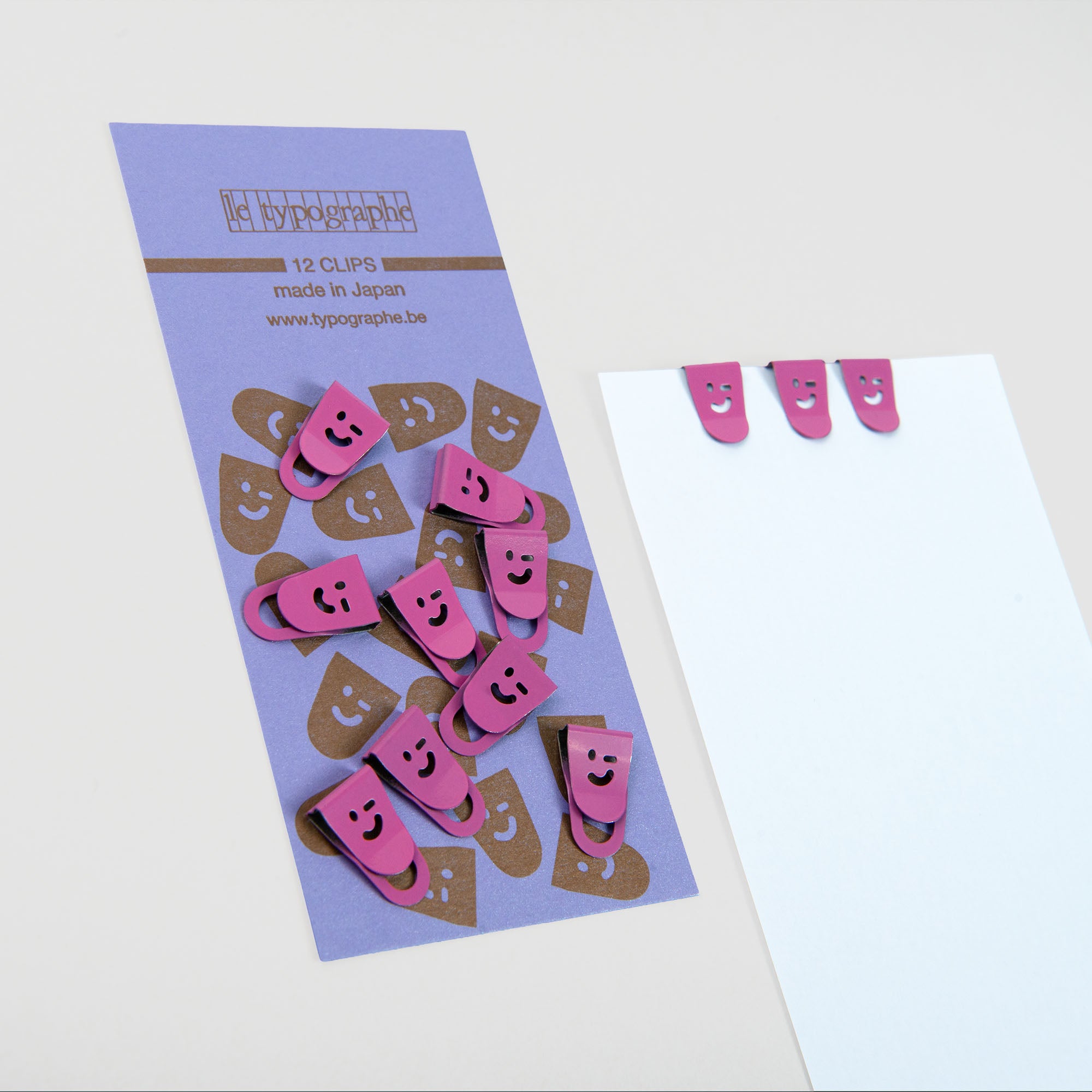 Mini Clips Smiley - Rosé von Le Typographe: ein Set mit 12 rosa Smiley-Mini-Clips, die auf einer hellvioletten Karte abgebildet sind, von denen drei auf einem weißen Blatt befestigt sind - perfekt für die Organisation auf dem Schreibtisch. Hergestellt in Japan.